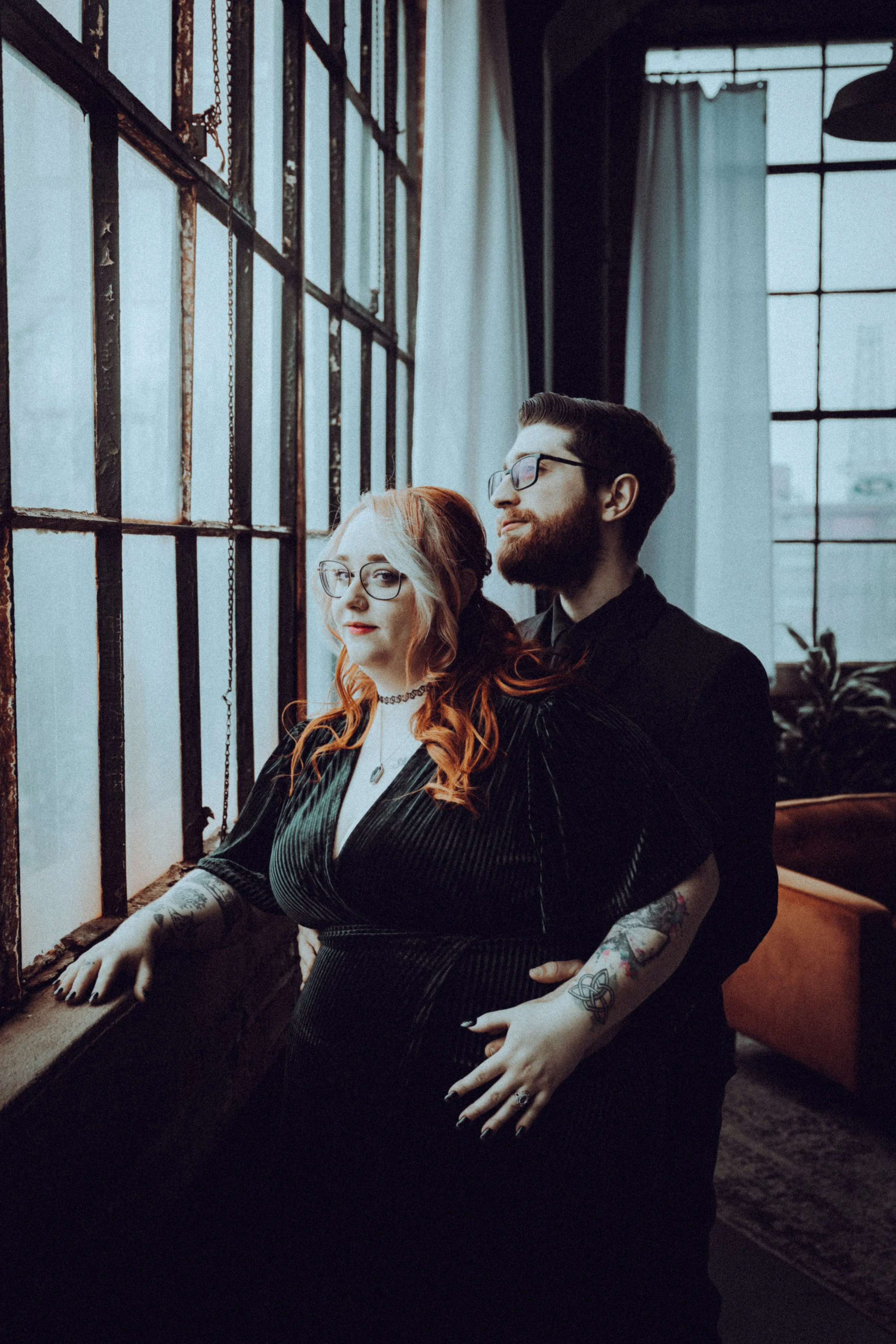 A couple standing near a large window with iron bars, in a dimly lit room with curtains and a couch in the background.