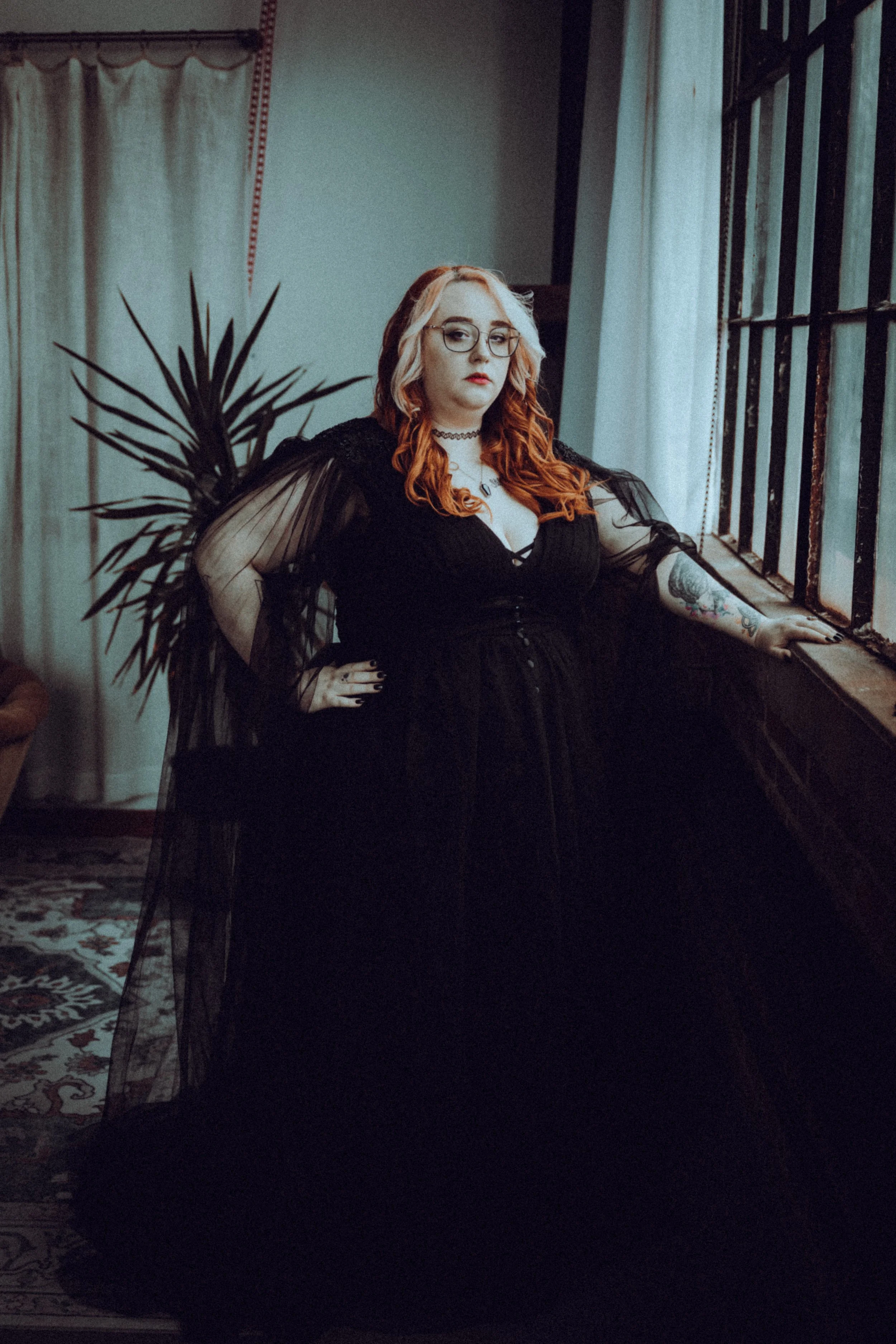 A woman with red curly hair, glasses, and tattoos wearing a black dress, standing by a large window with her hand resting on the windowsill, in a room with curtains and patterned carpet, looking at the camera.