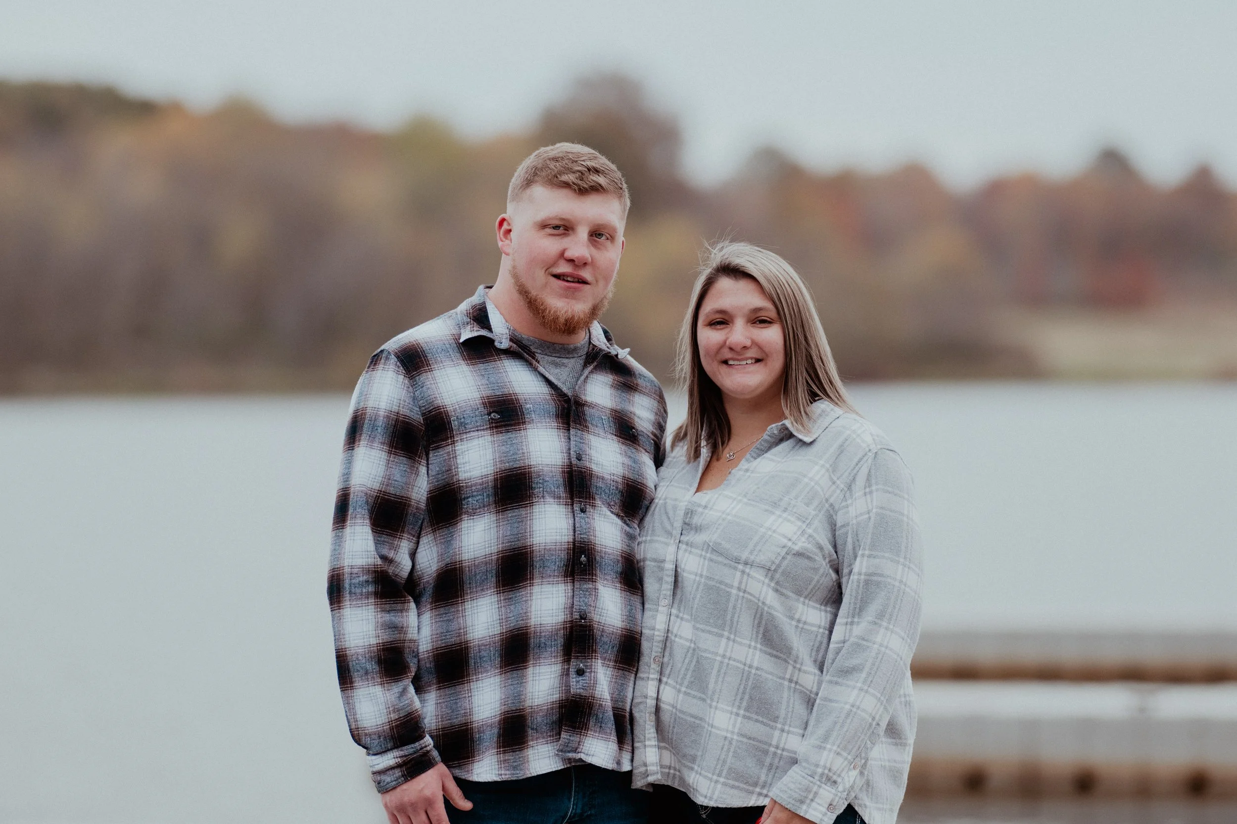A young man and woman stand together outdoors near a body of water, both smiling and wearing casual plaid shirts, with a blurred background of trees and water.