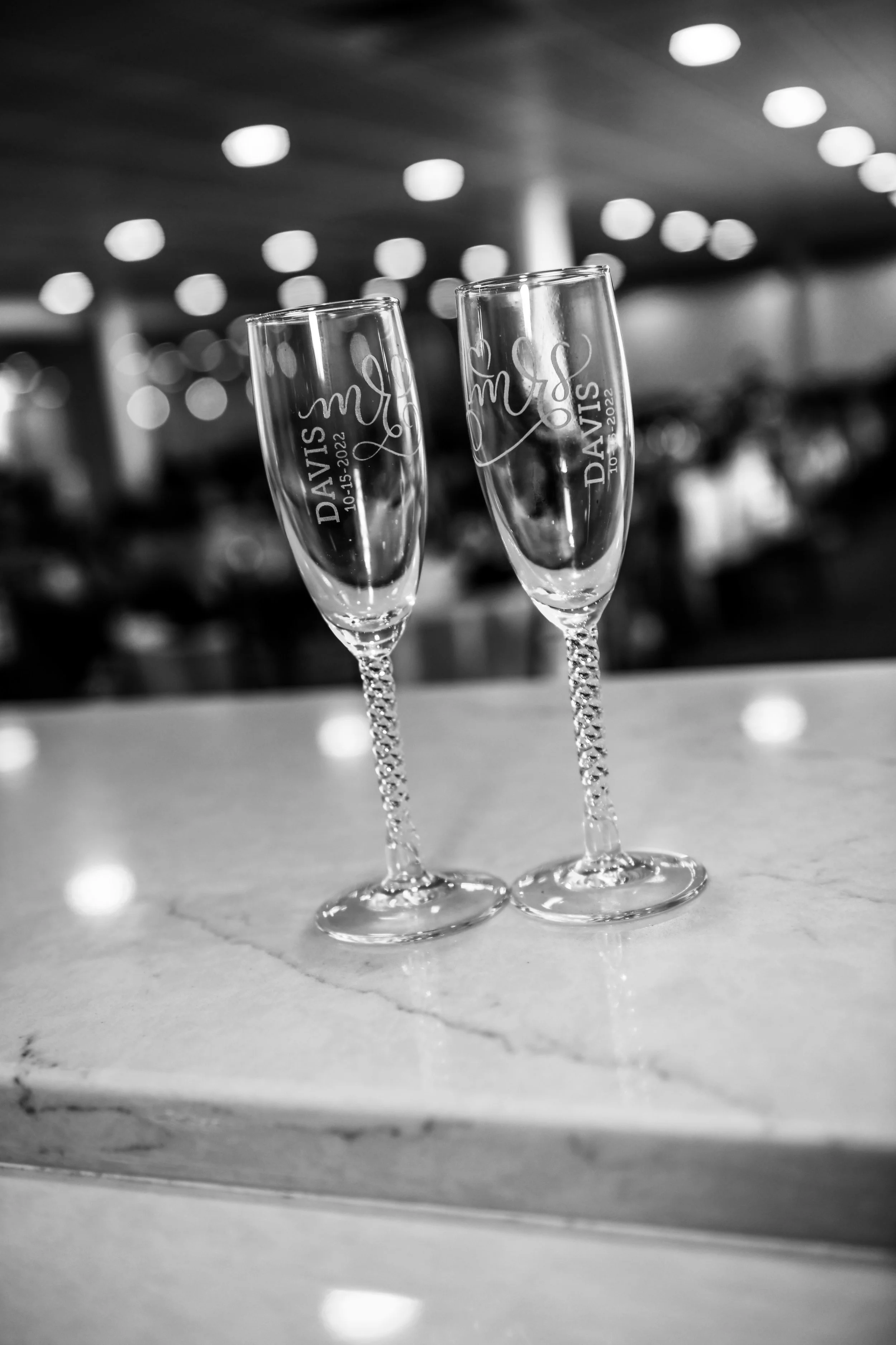 Two engraved champagne flutes with twisted stems on a marble surface, blurred background with circular ceiling lights.
