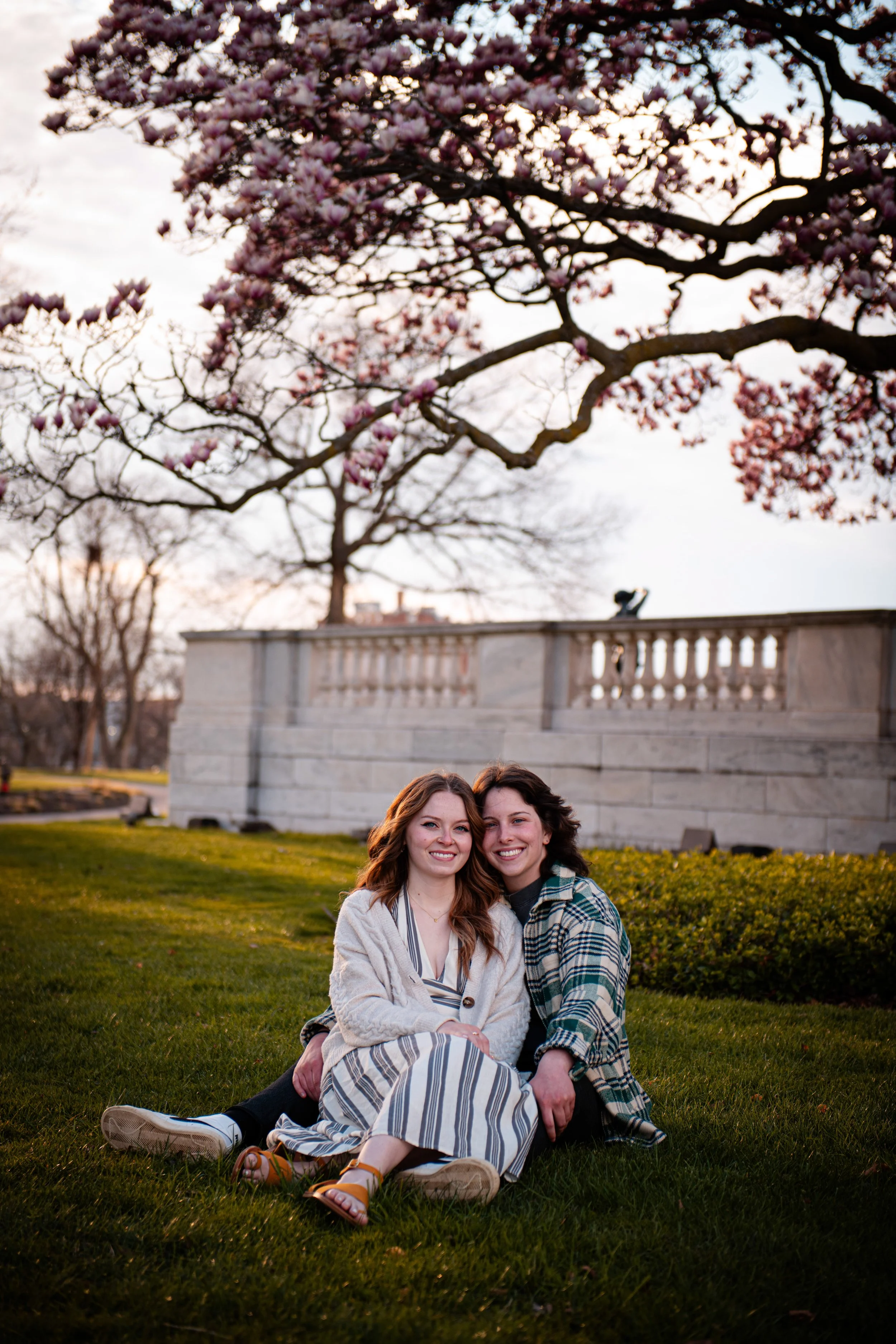 Two young women sitting on grass in a park, smiling and leaning close together, with blooming pink cherry blossom trees and a stone structure in the background during sunset.