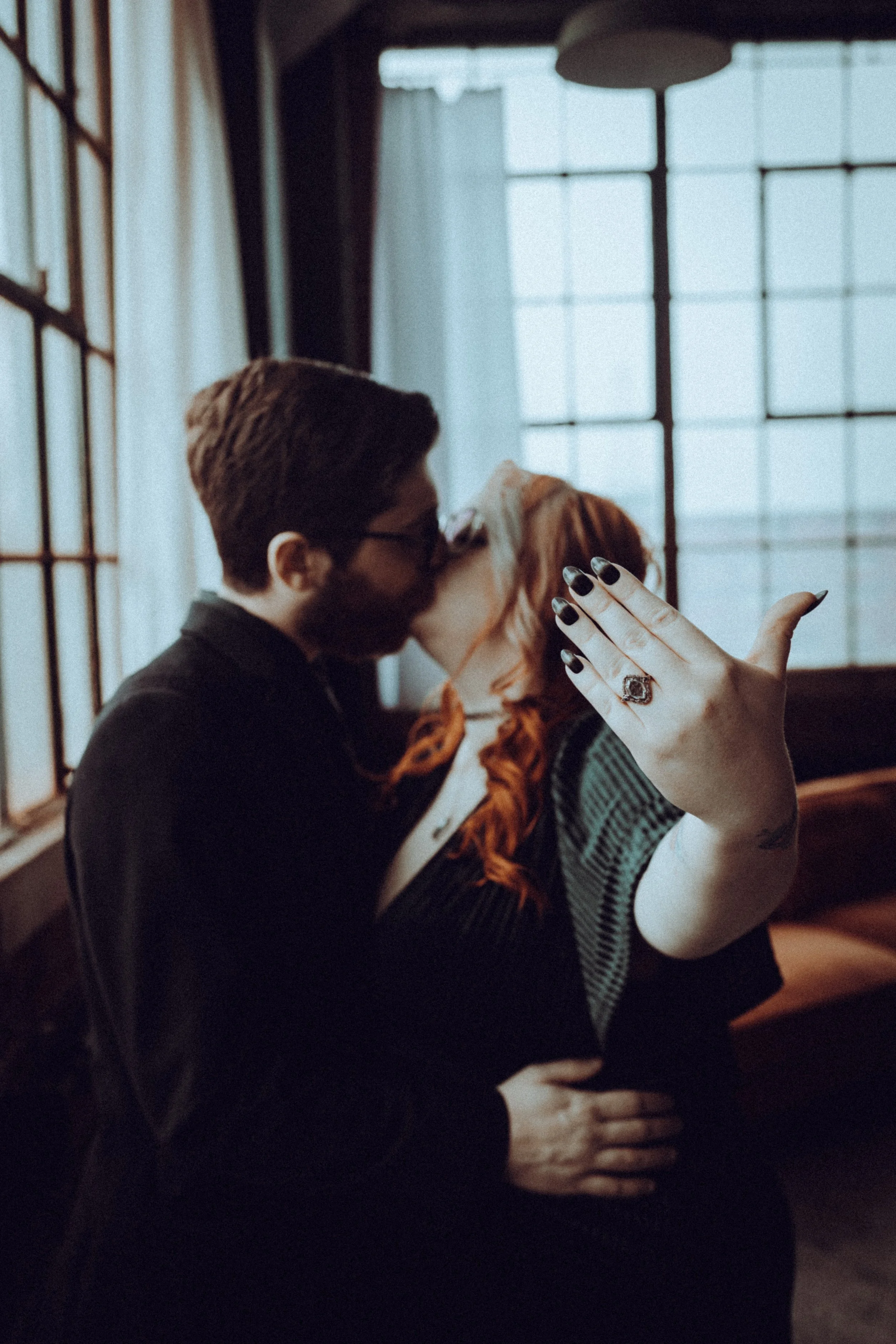 A couple kissing in a dimly lit room, with the woman showing off a black ring on her finger.