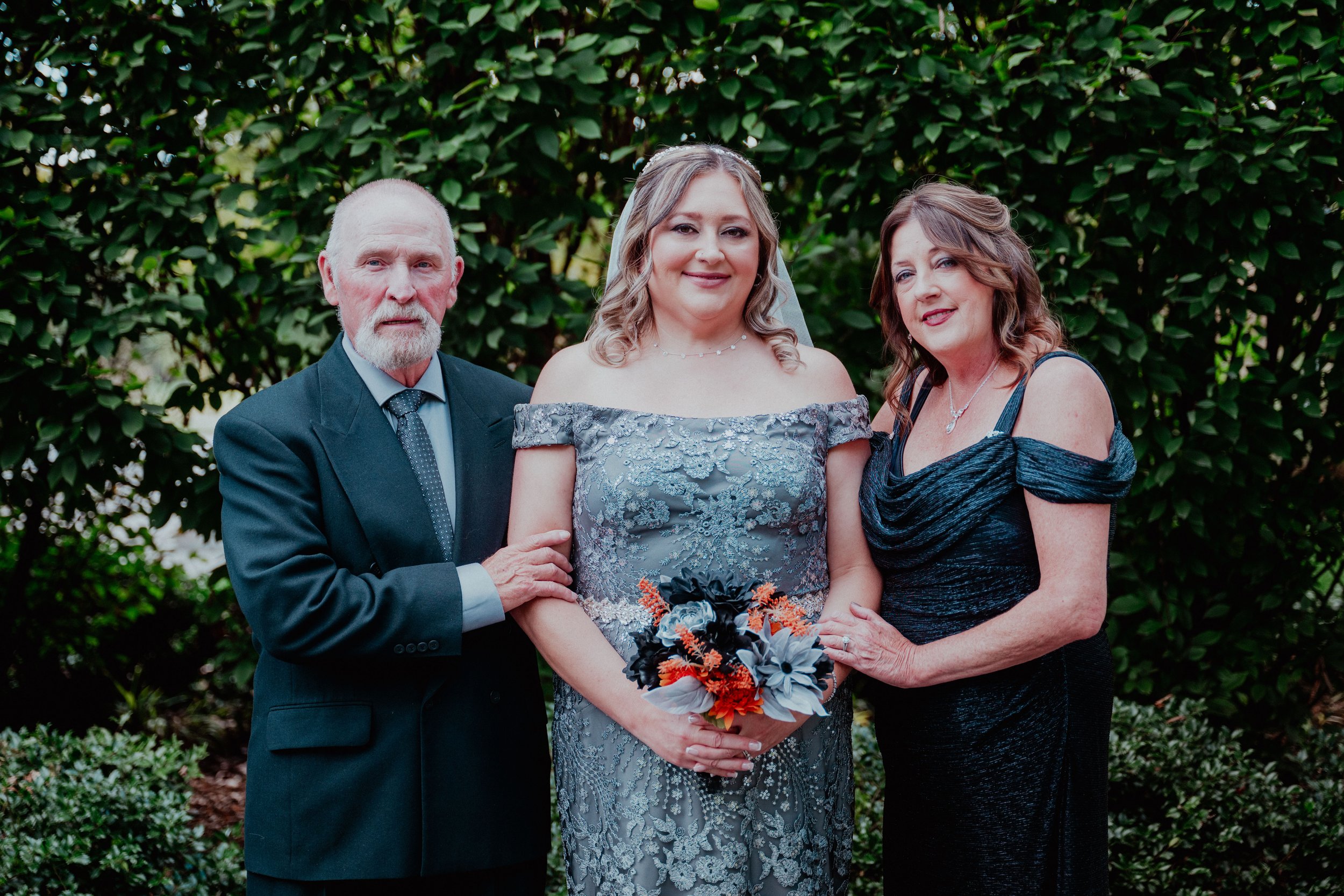 A woman in a silver lace dress holding a bouquet, standing between an older man in a dark suit and a woman in a dark off-shoulder dress, all posing outdoors in front of green foliage.
