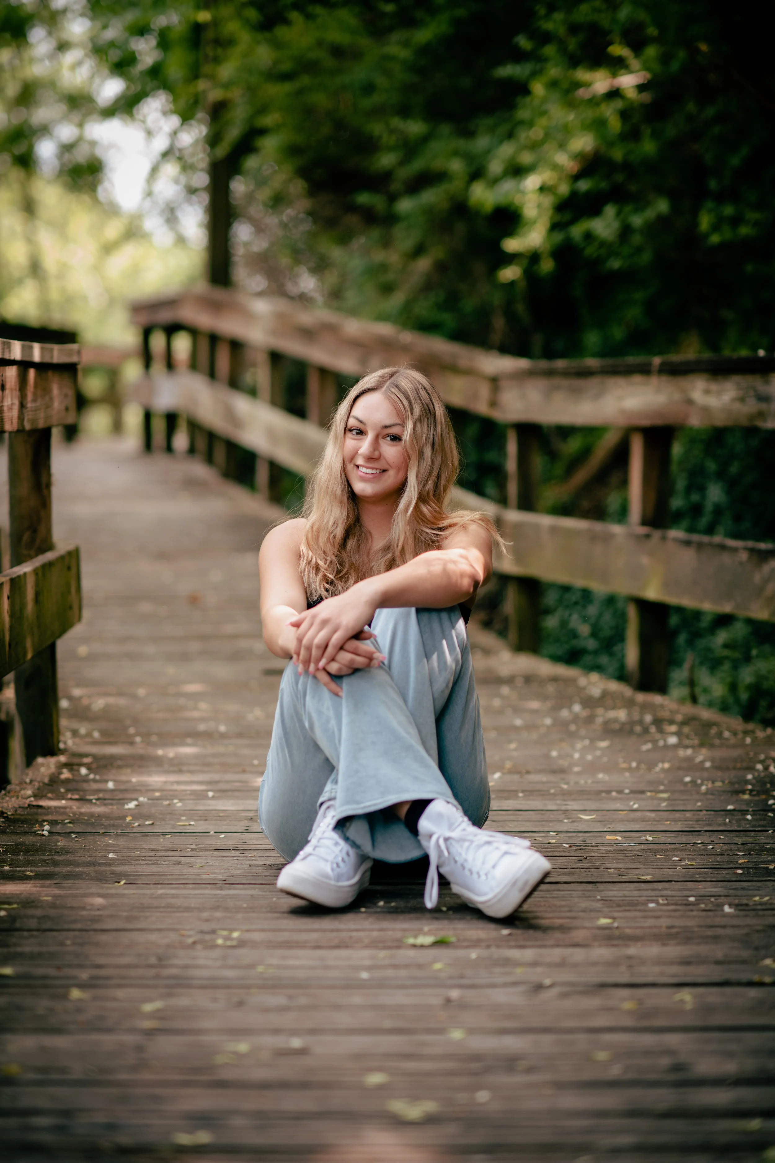 A young woman with long blonde hair, casual clothing, white sneakers, sitting on a wooden bridge in a forested area, smiling and relaxed.