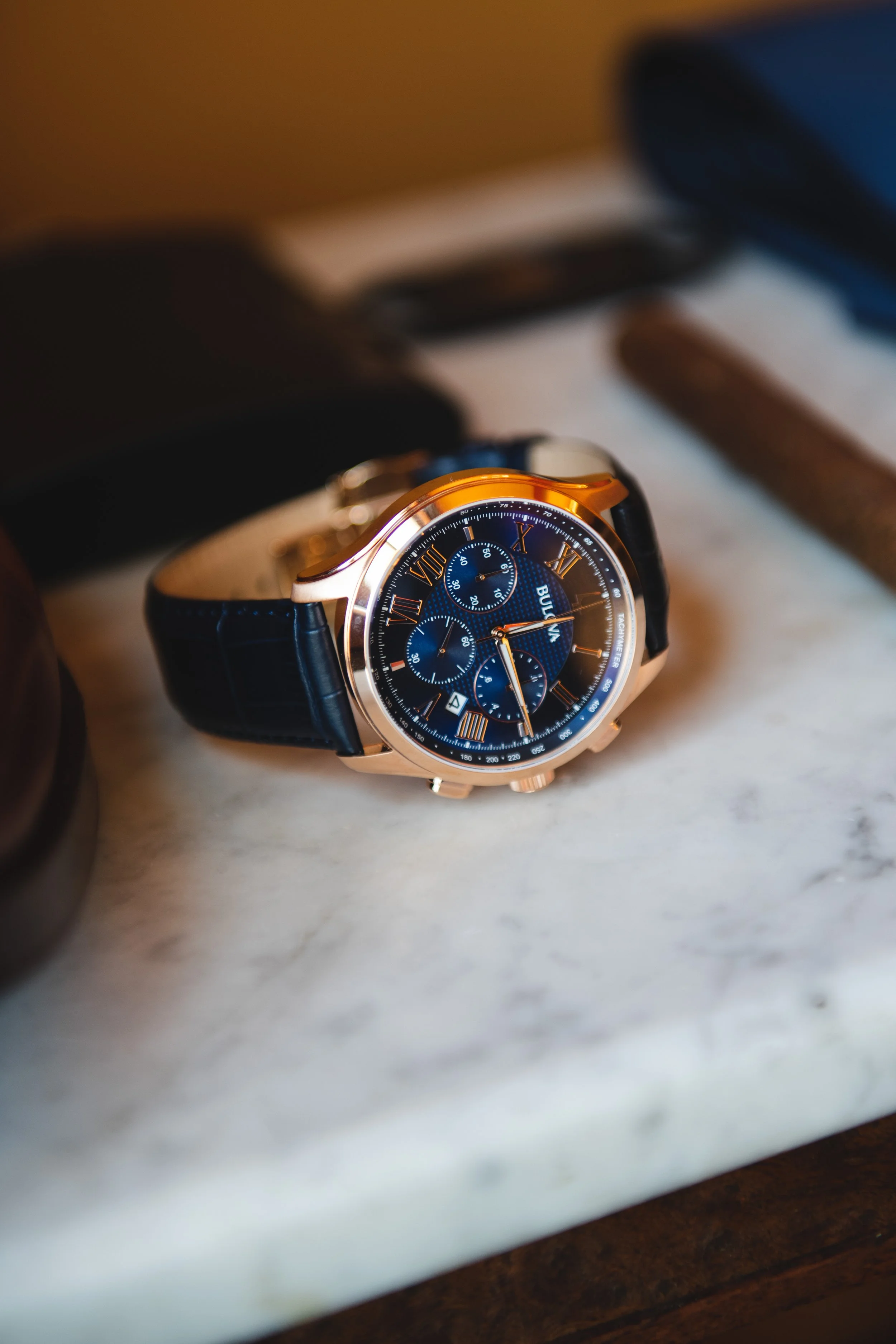 A luxury wristwatch with a blue dial, gold case, and leather strap resting on a marble surface, with blurred accessories in the background.