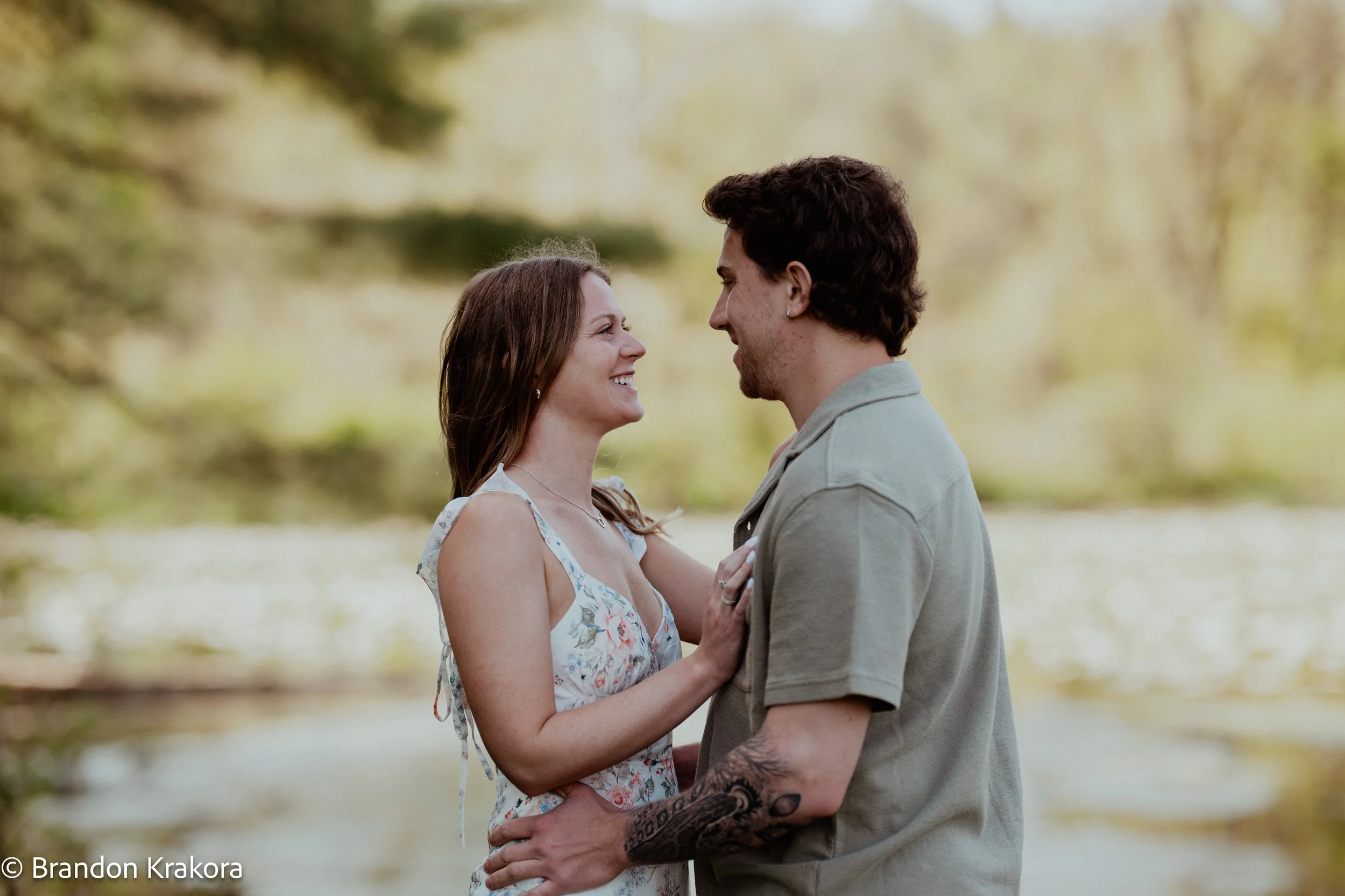 A couple standing close together outdoors near a body of water, smiling at each other, with trees in the background.