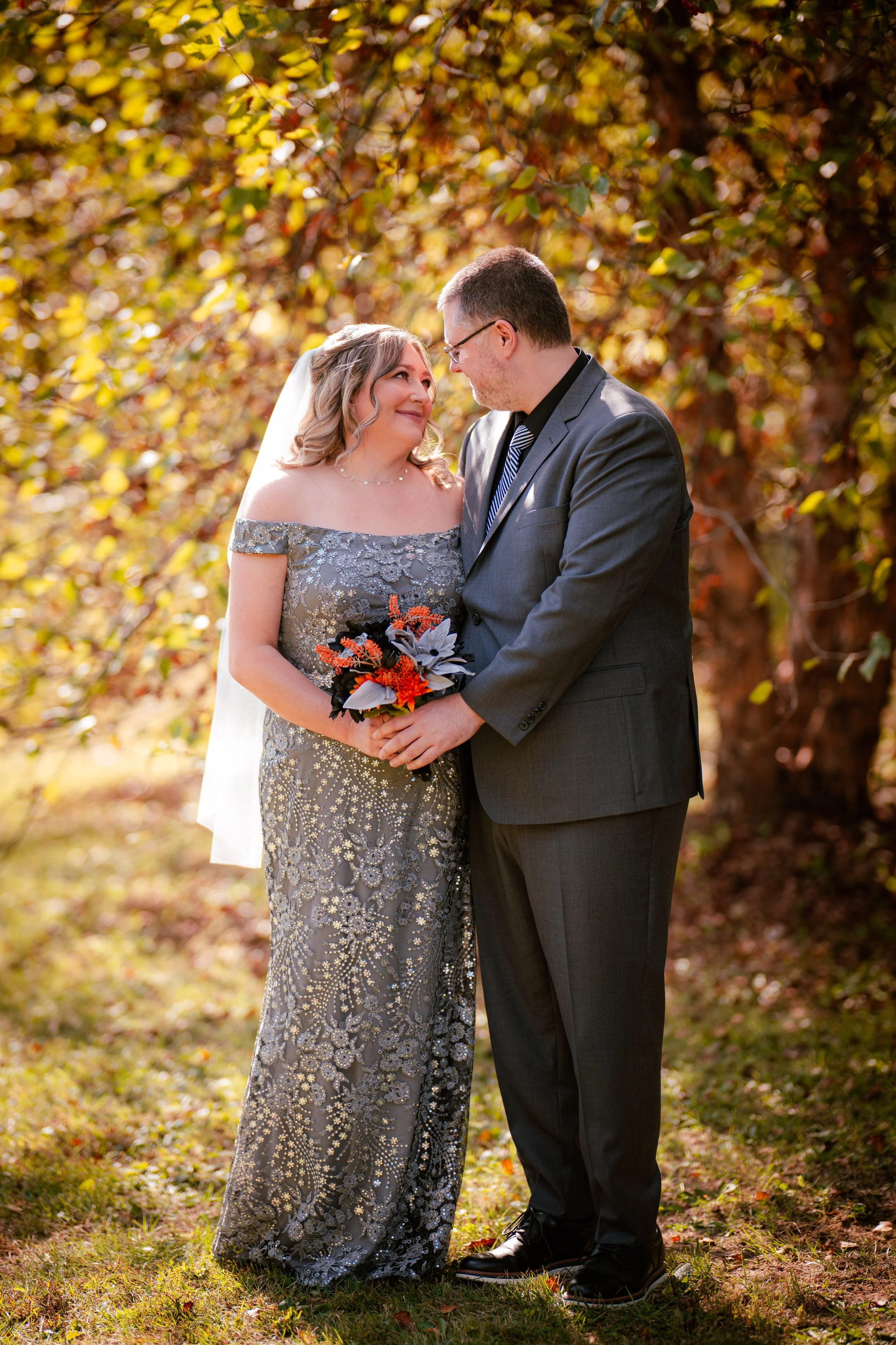 A bride and groom standing outdoors, gazing into each other's eyes, with autumn foliage in the background. The bride wears an off-the-shoulder, silver embellished gown and holds a bouquet of orange, black, and white flowers. The groom is dressed in a