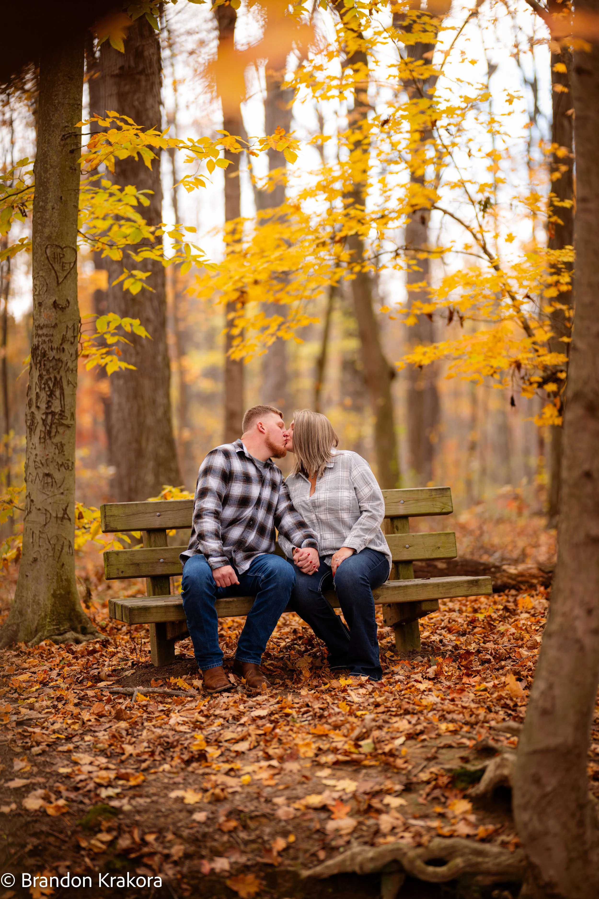 A couple sitting on a wooden park bench in a forest with autumn leaves, kissing and holding hands.
