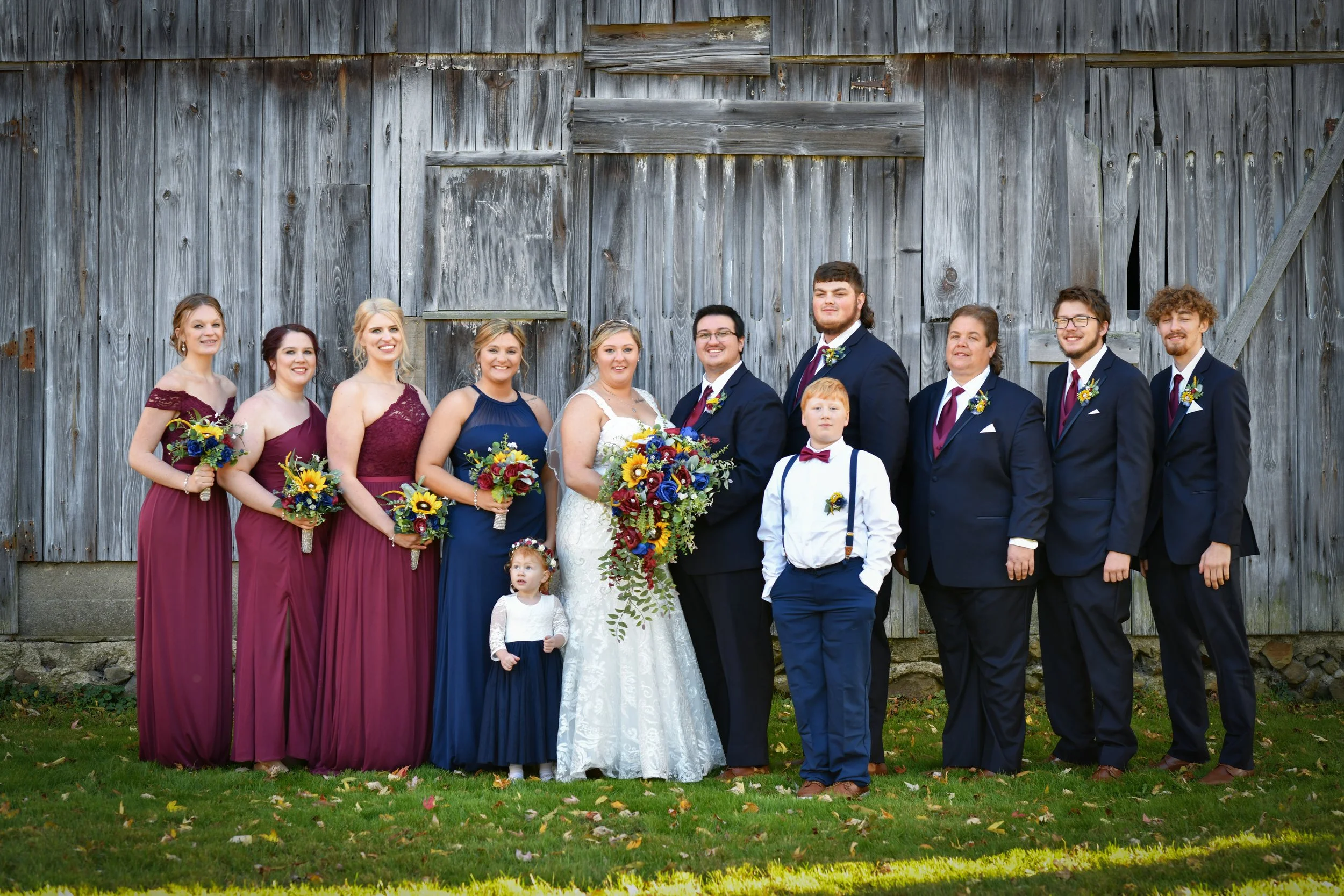 A wedding party standing in front of a wooden barn. The group includes a bride in a white wedding dress, a groom in a dark suit, six bridesmaids in burgundy and navy dresses, two young boys in formal attire, and a flower girl. They are holding bouque