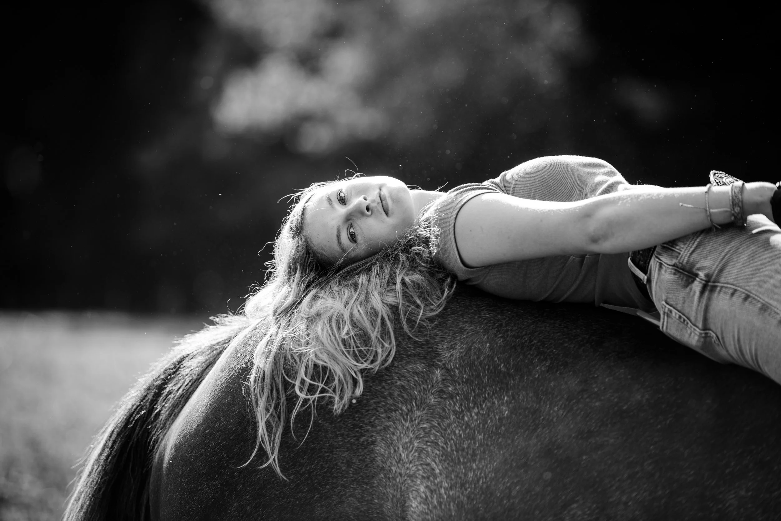 A woman with wavy hair lying on her side on a horse in a natural outdoor setting, black and white photography.