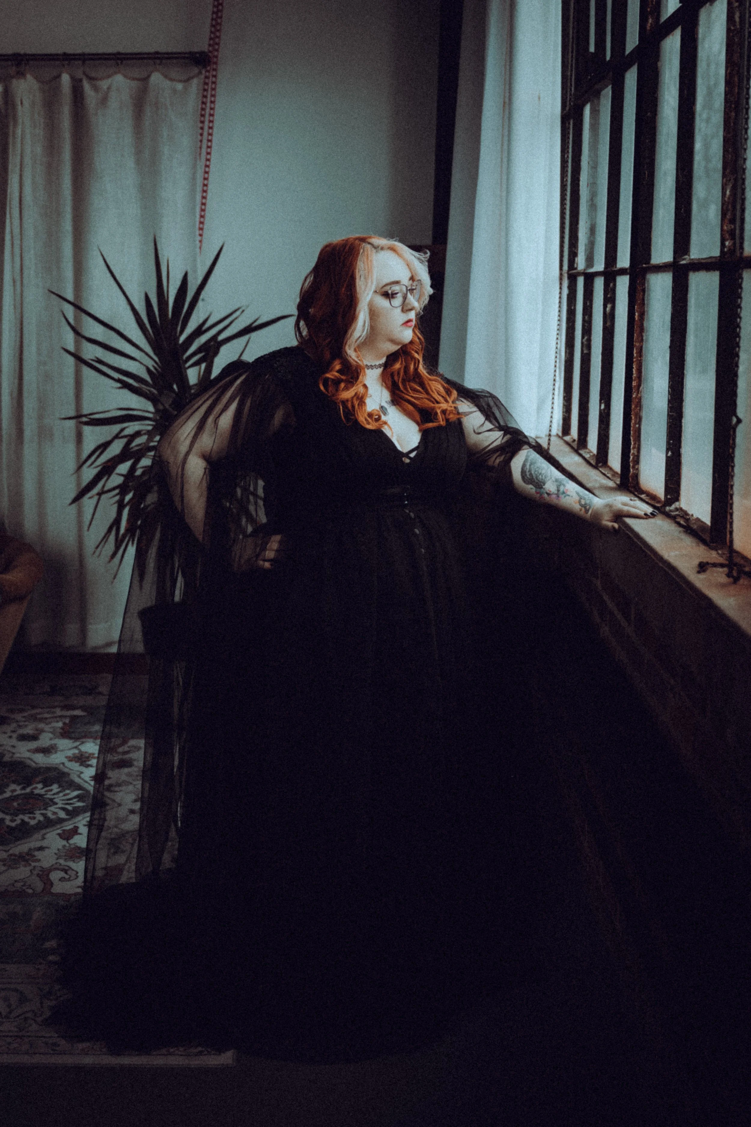 A woman with red hair and glasses wearing a black dress with sheer sleeves, sitting on a windowsill in a dimly lit room.