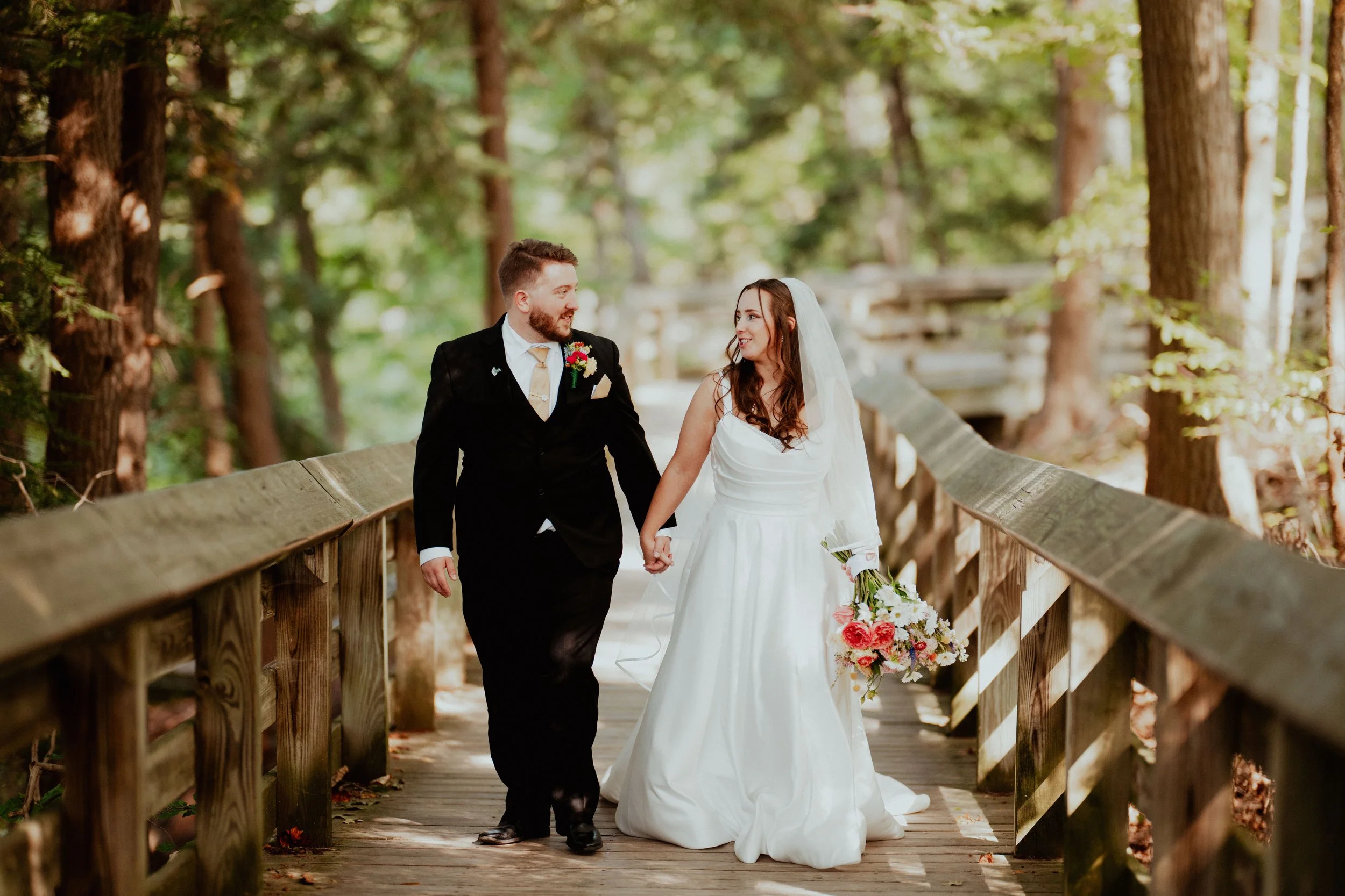 A bride and groom walking hand in hand on a wooden bridge surrounded by trees, during their wedding day, with sunlight filtering through the leaves.