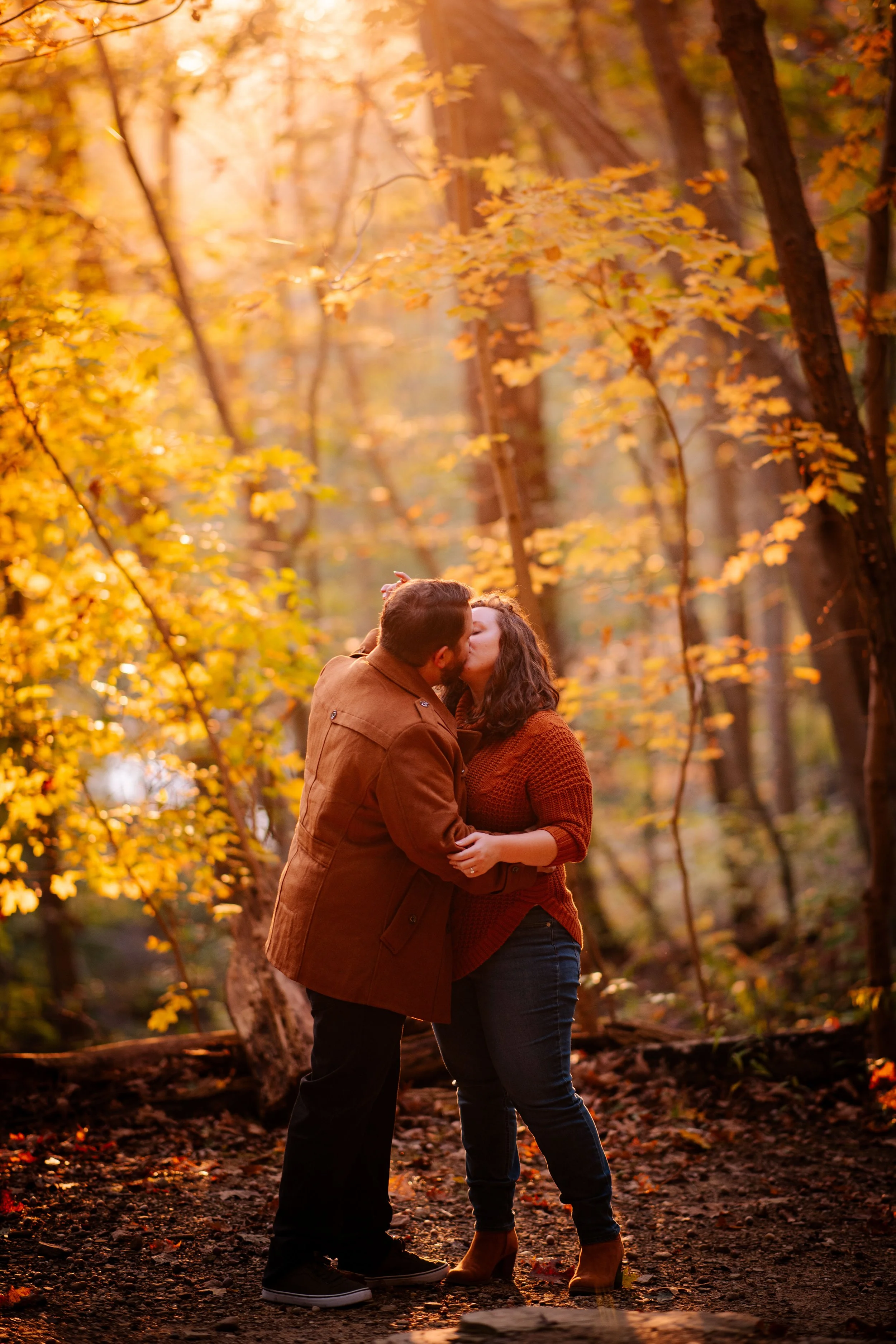 A couple kissing in a forest during autumn, surrounded by yellow and orange leaves on trees, with sunlight filtering through the branches.