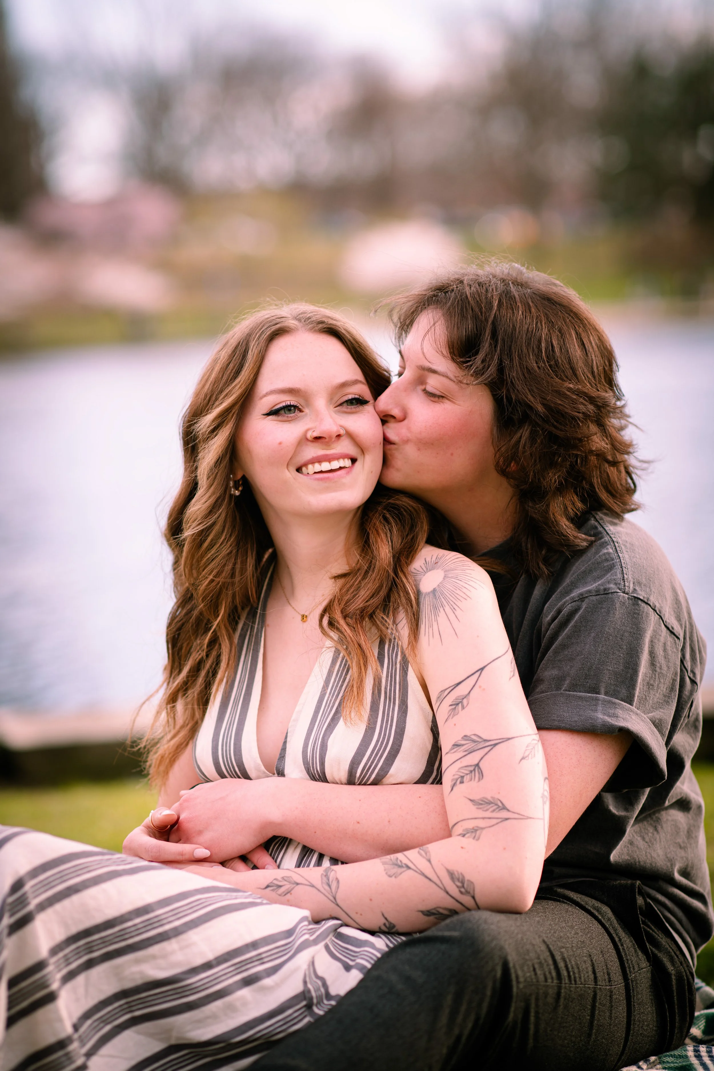 A young couple sitting outdoors near a body of water, with the man kissing the woman on the cheek, both smiling, with cherry blossoms in the background.