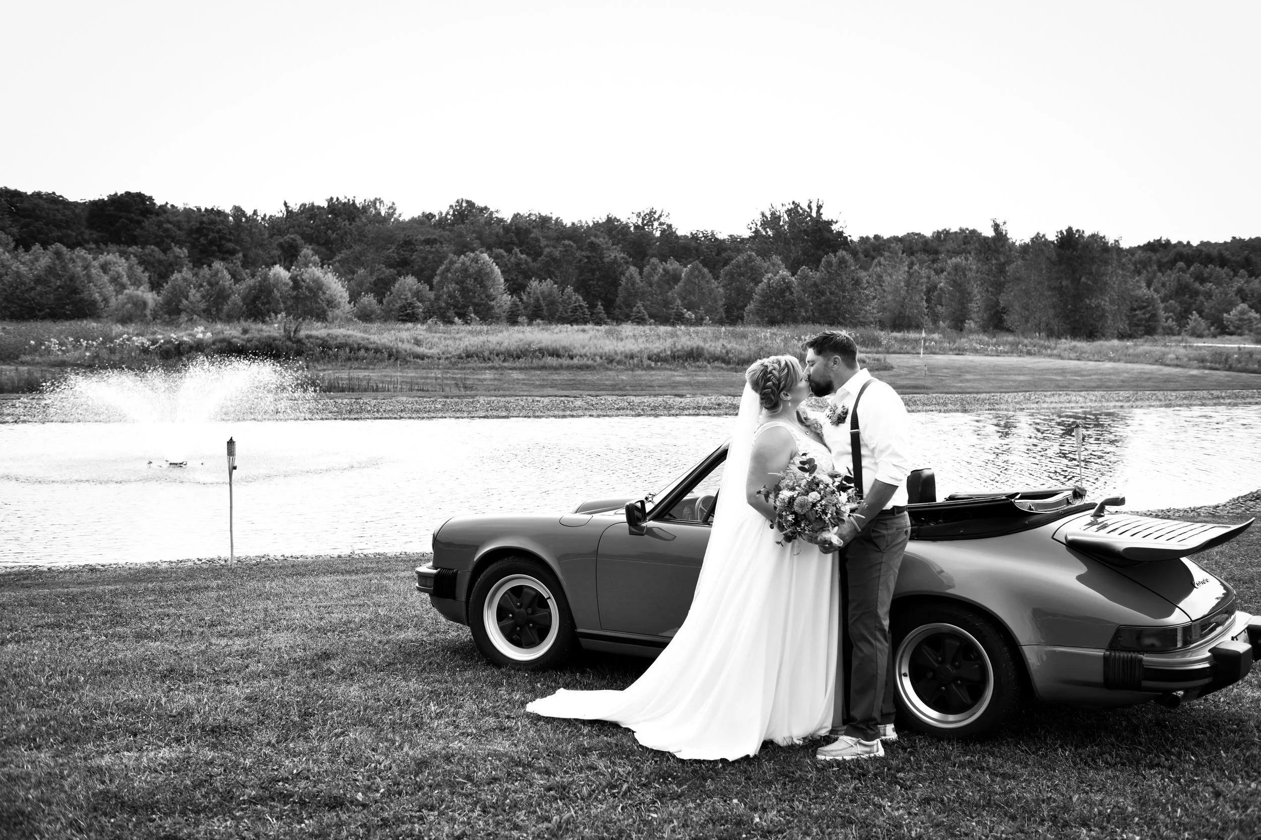 Black and white photo of a bride and groom kissing beside a vintage car near a lake, with trees in the background.