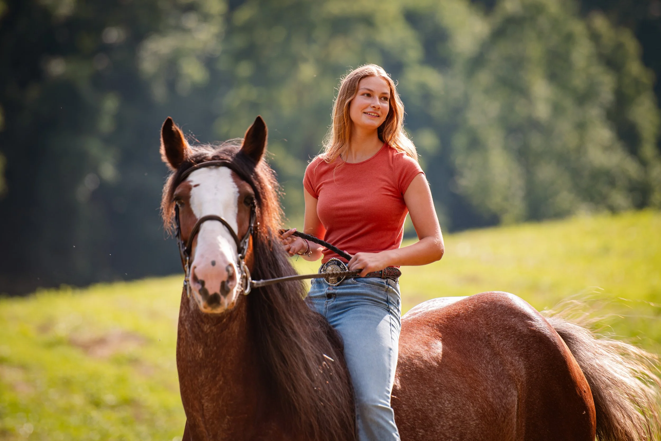 A woman riding a brown and white horse in a grassy field with trees in the background.