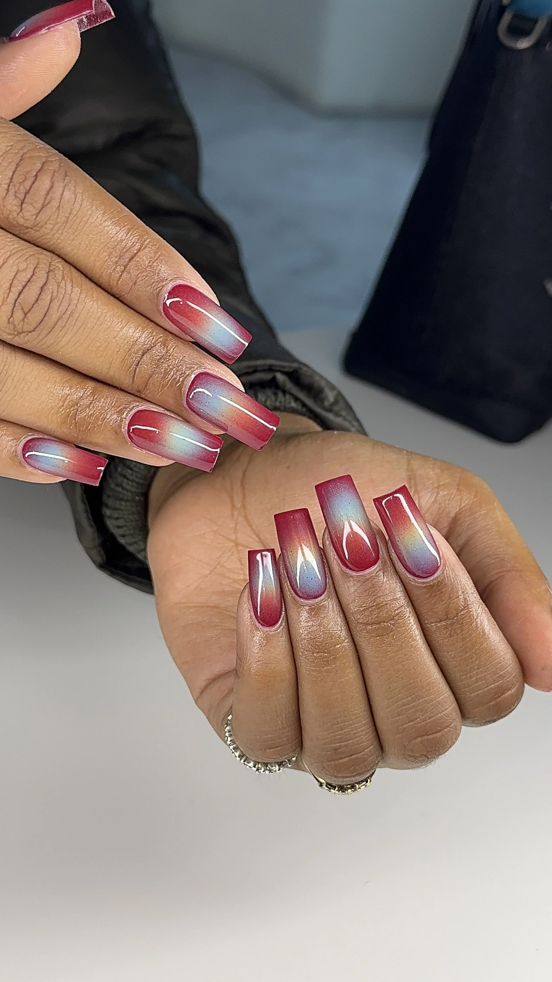 A person's hand with long, square-shaped nails displaying a colorful gradient nail art in red, pink, purple, yellow, and blue hues.