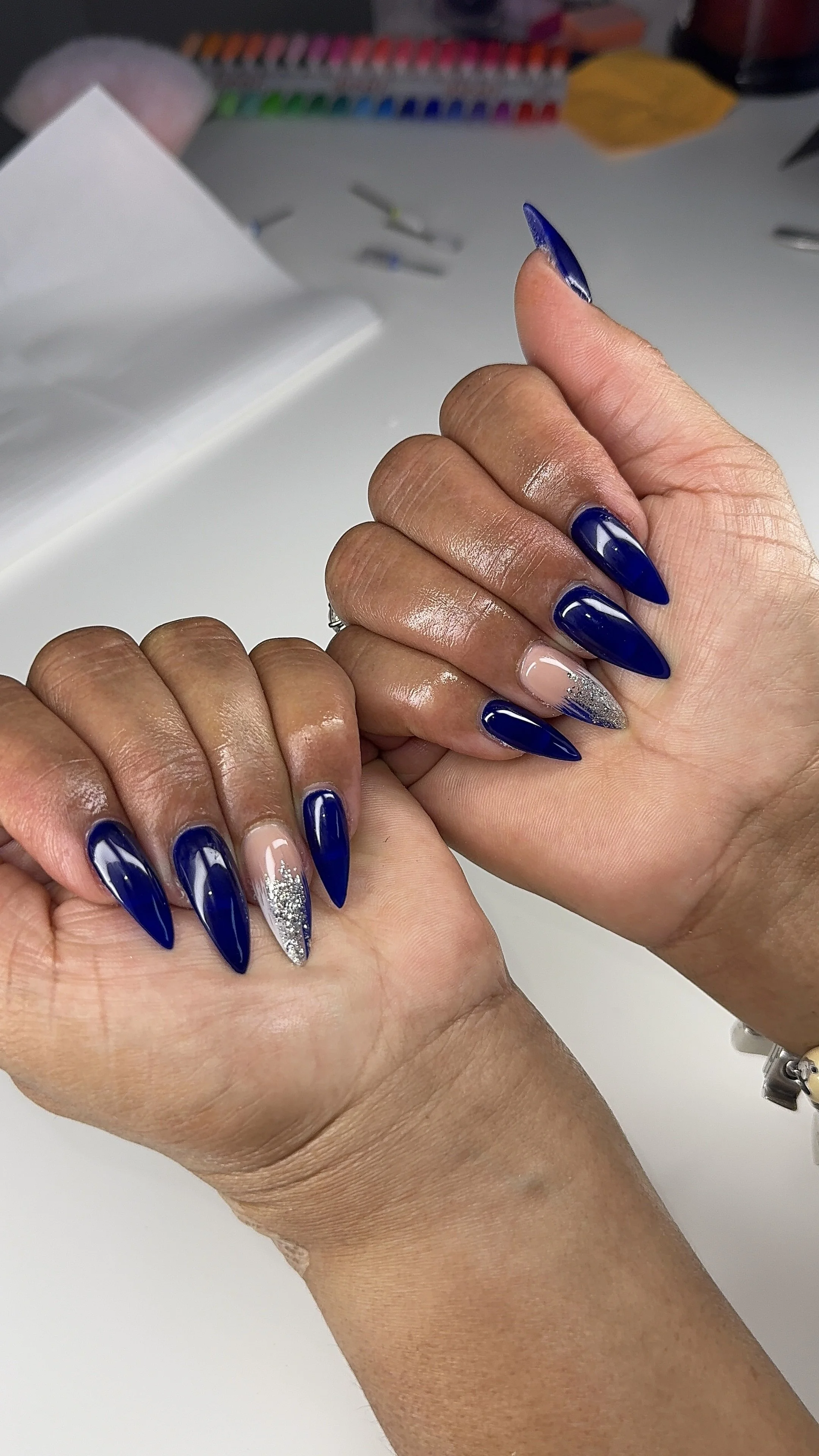 Close-up of hands with long, almond-shaped, dark blue and silver glitter acrylic nails, on a white surface.