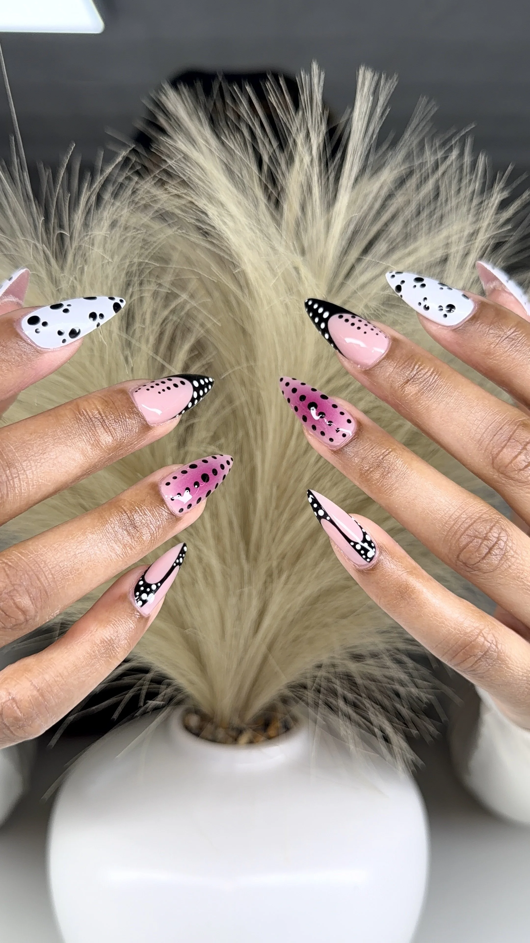 Close-up of hands with long, decorated nails with pink, white, and black polka dot and stripe designs, positioned around a white vase with dried beige pampas grass.