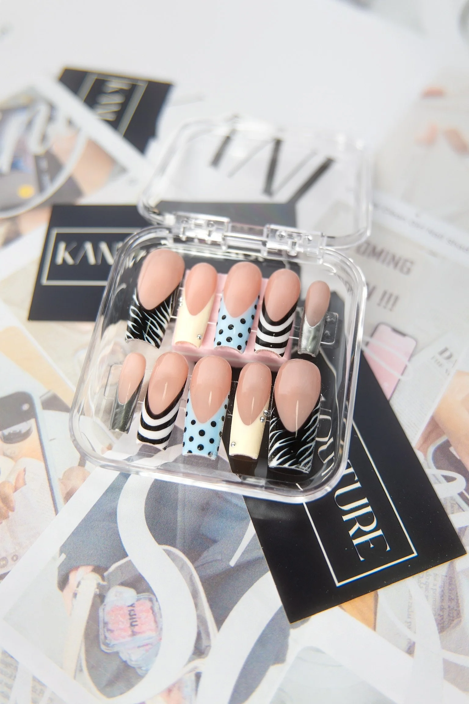 A set of decorative fake nails in a clear plastic case, with black, white, light blue, and beige designs, placed on a magazine with fashion advertisements.