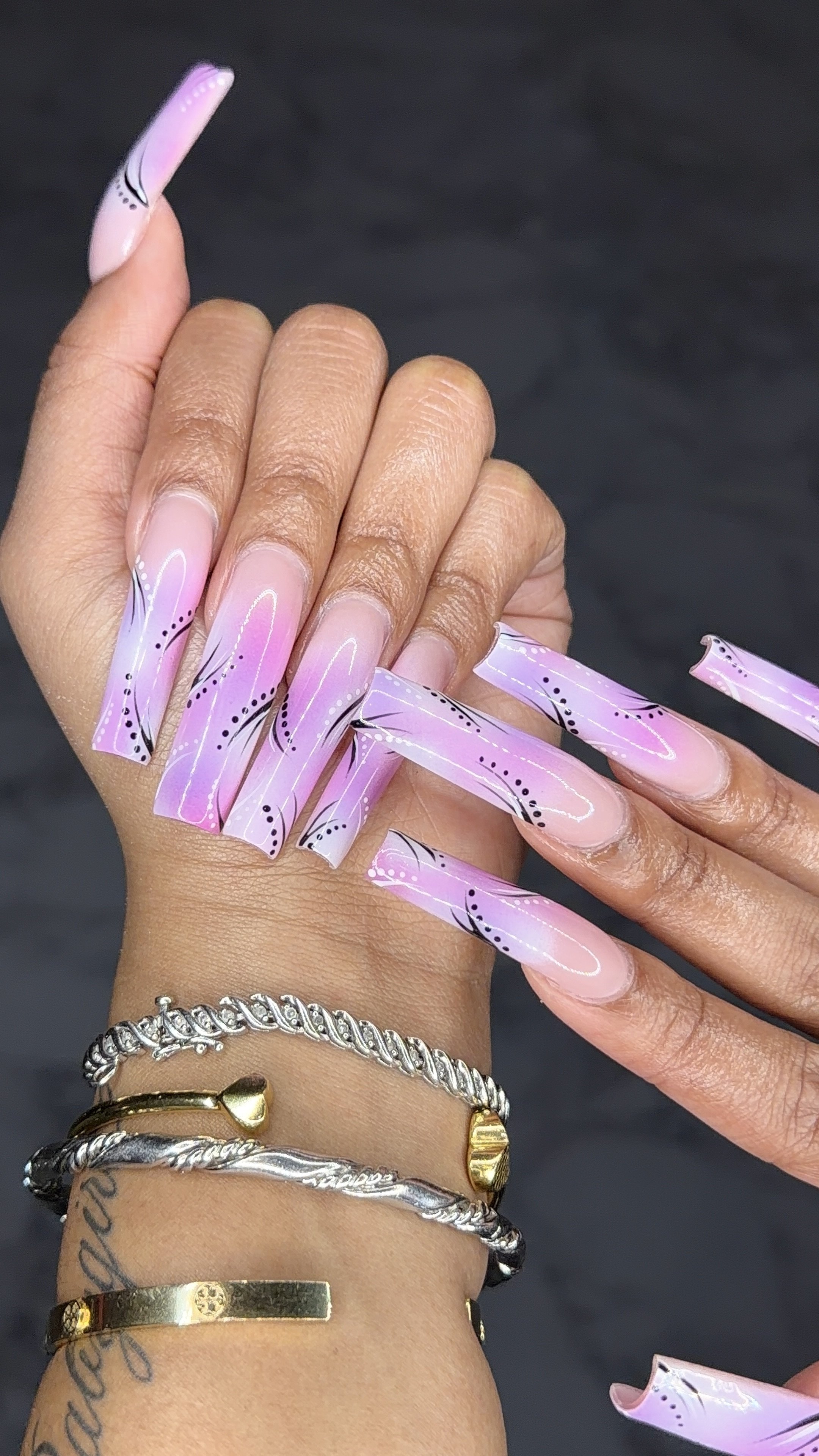 Close-up of a hand with long, pink, and black painted acrylic nails and gold and silver bracelets on the wrist.