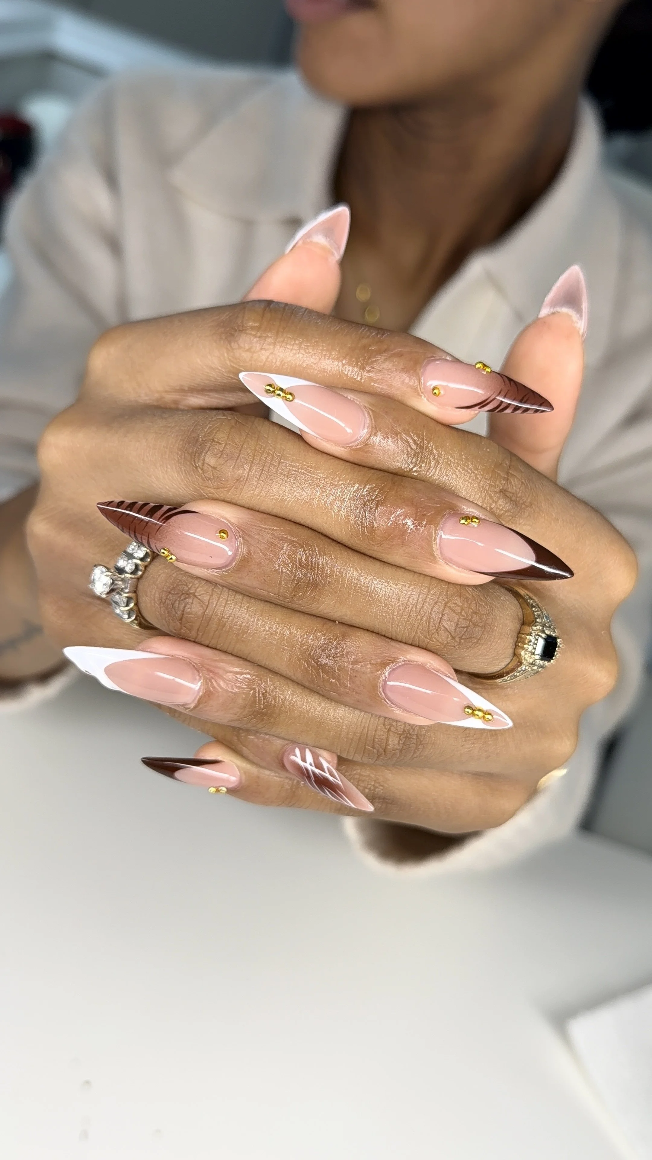 Close-up of a woman's hands with long, almond-shaped nails featuring a pink, white, and brown design, gold accents, and decorative patterns.