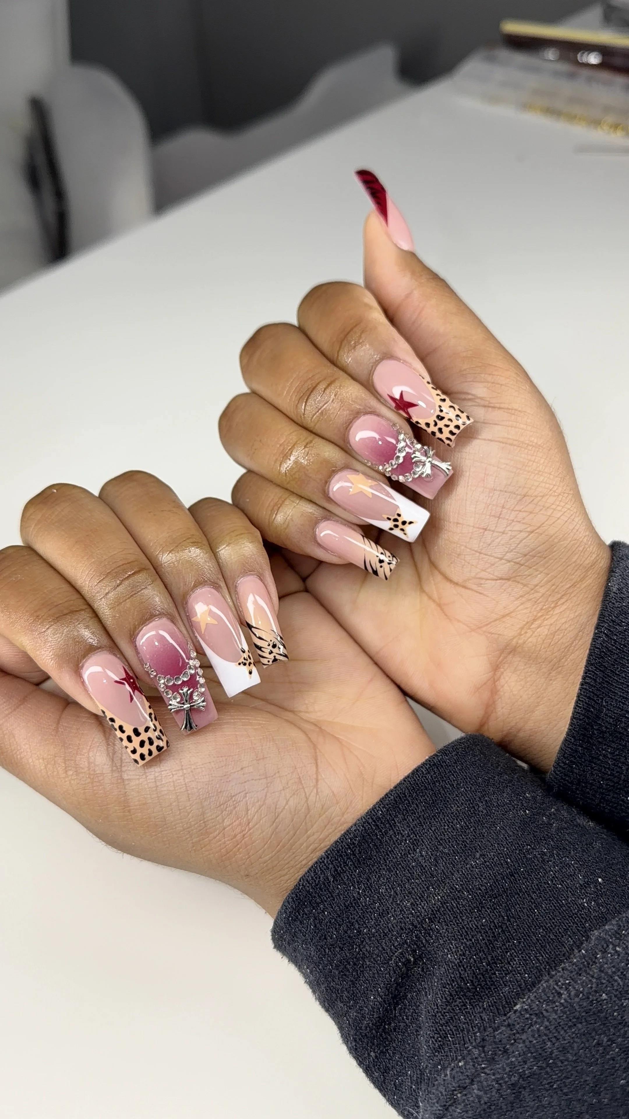 Close-up of hands with elaborate nail art featuring pink, white, and nude colors with animal print, stars, and red star designs, accented with rhinestones and jewelry.
