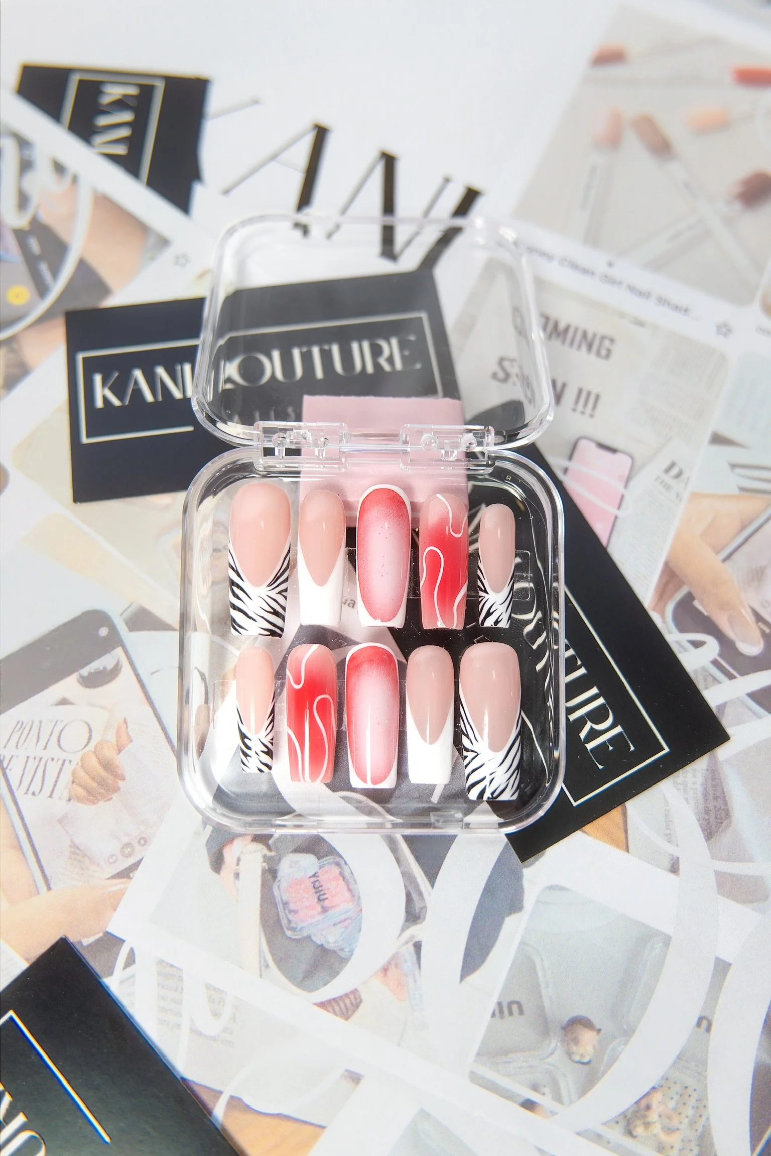 A clear plastic case with decorative nail tips inside, placed on a magazine. The nail tips have designs including pink, white, black and white zebra patterns, and pinkish-red with white swirl details.