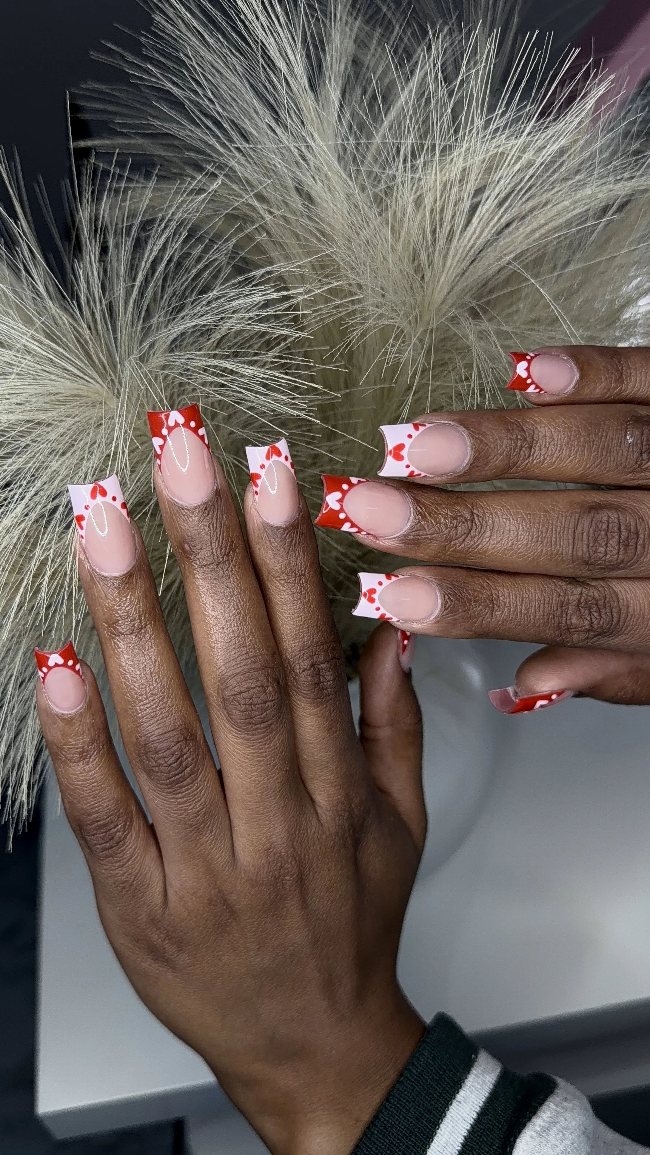 Close-up of hands with manicured nails featuring a red and white heart pattern, positioned near fluffy, cream-colored dried grasses.