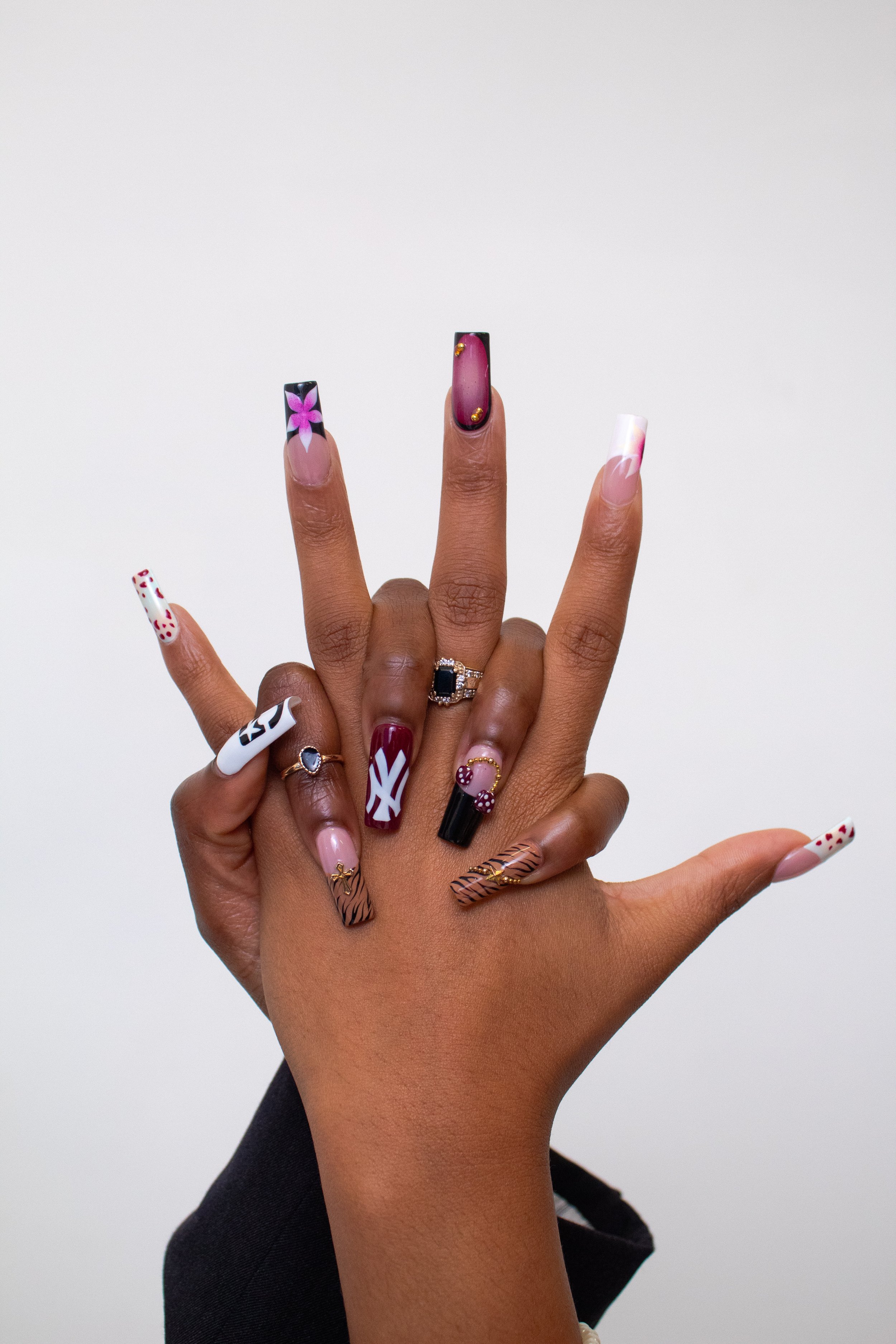 Close-up of two hands with decorated nails and multiple rings. The nails feature various colorful and intricate designs, including flowers, stripes, and abstract patterns, against a plain white background.