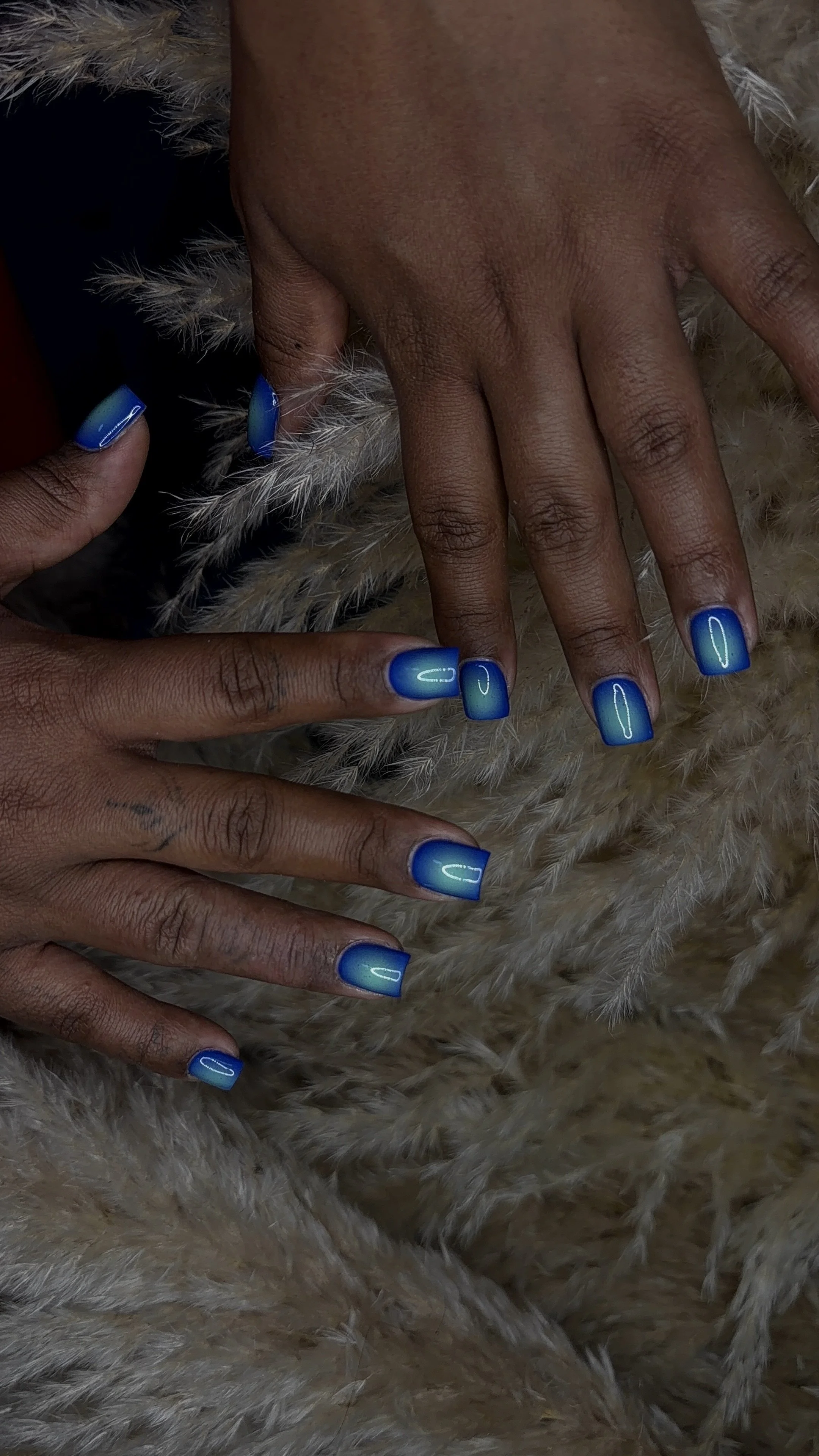 A person with dark skin showing off their blue ombre nails, resting on a soft, furry surface.