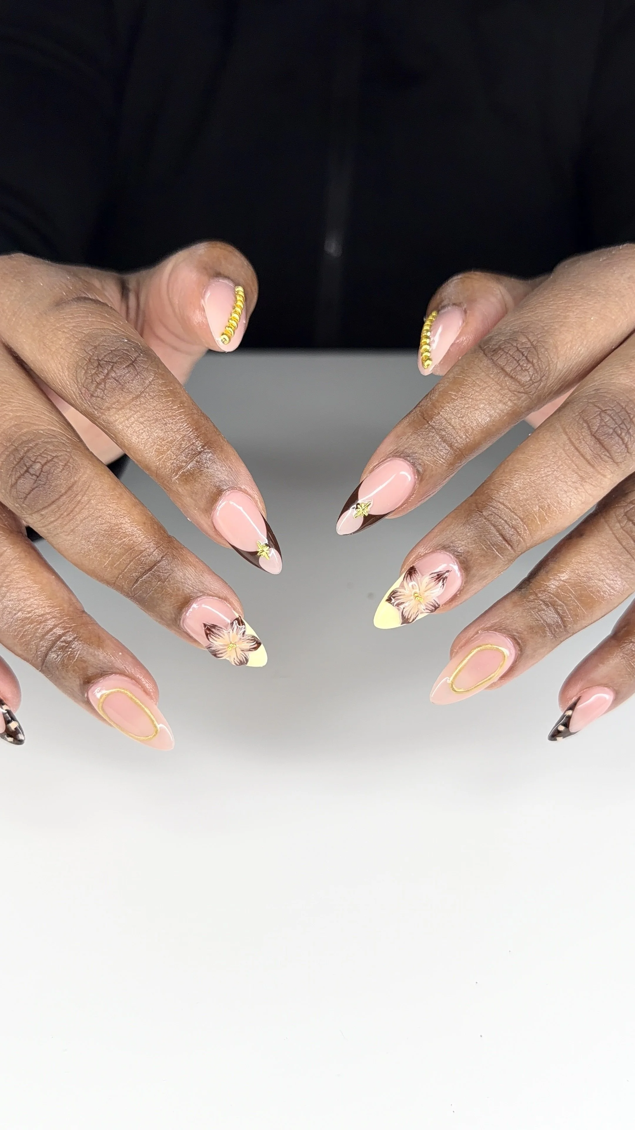 Close-up of hands with manicured nails featuring flower and abstract designs in pink, yellow, and black colors, with decorative gold rings on some fingers, against a neutral background.