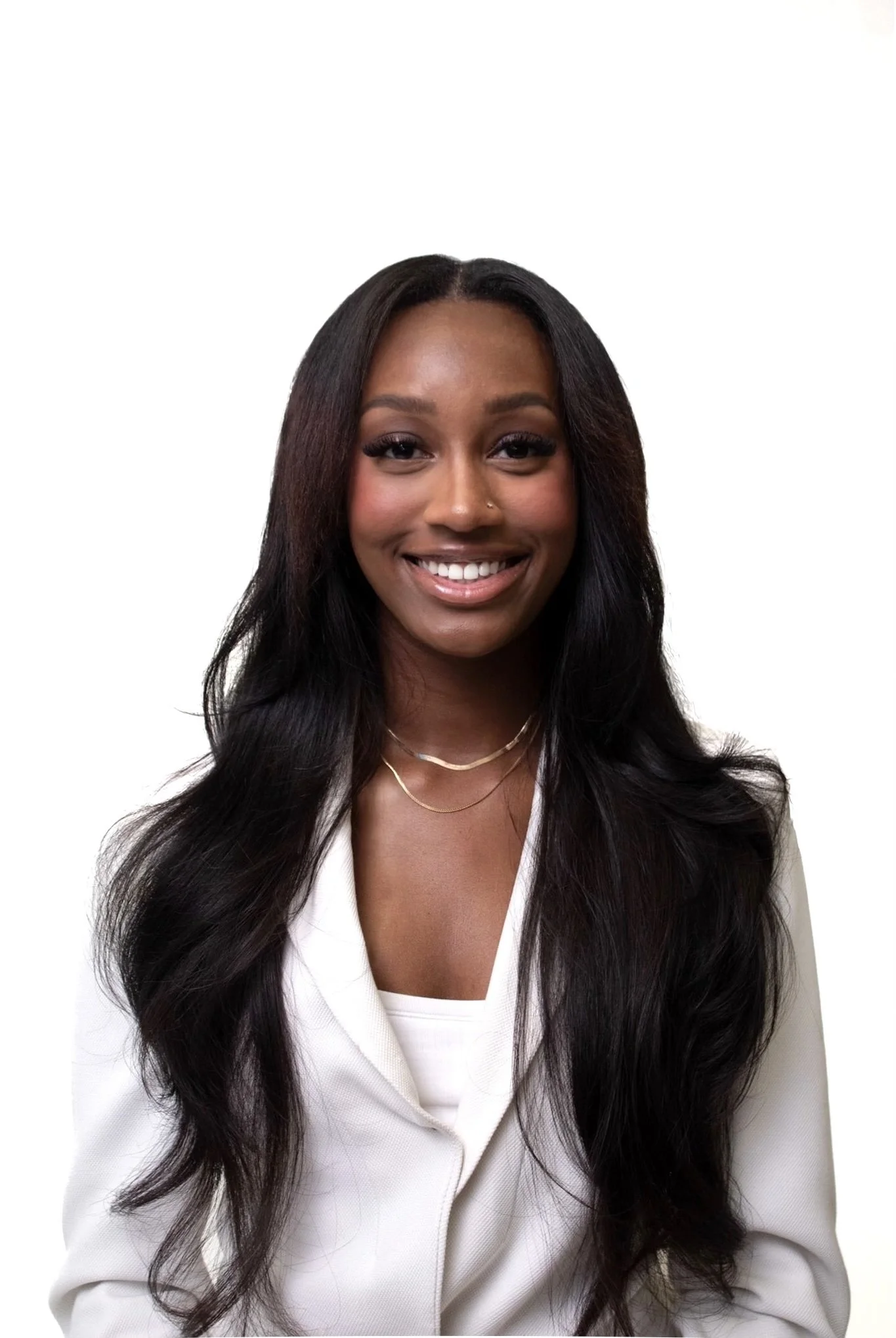 A smiling woman with long black wavy hair wearing a white blazer and layered necklaces against a plain white background.