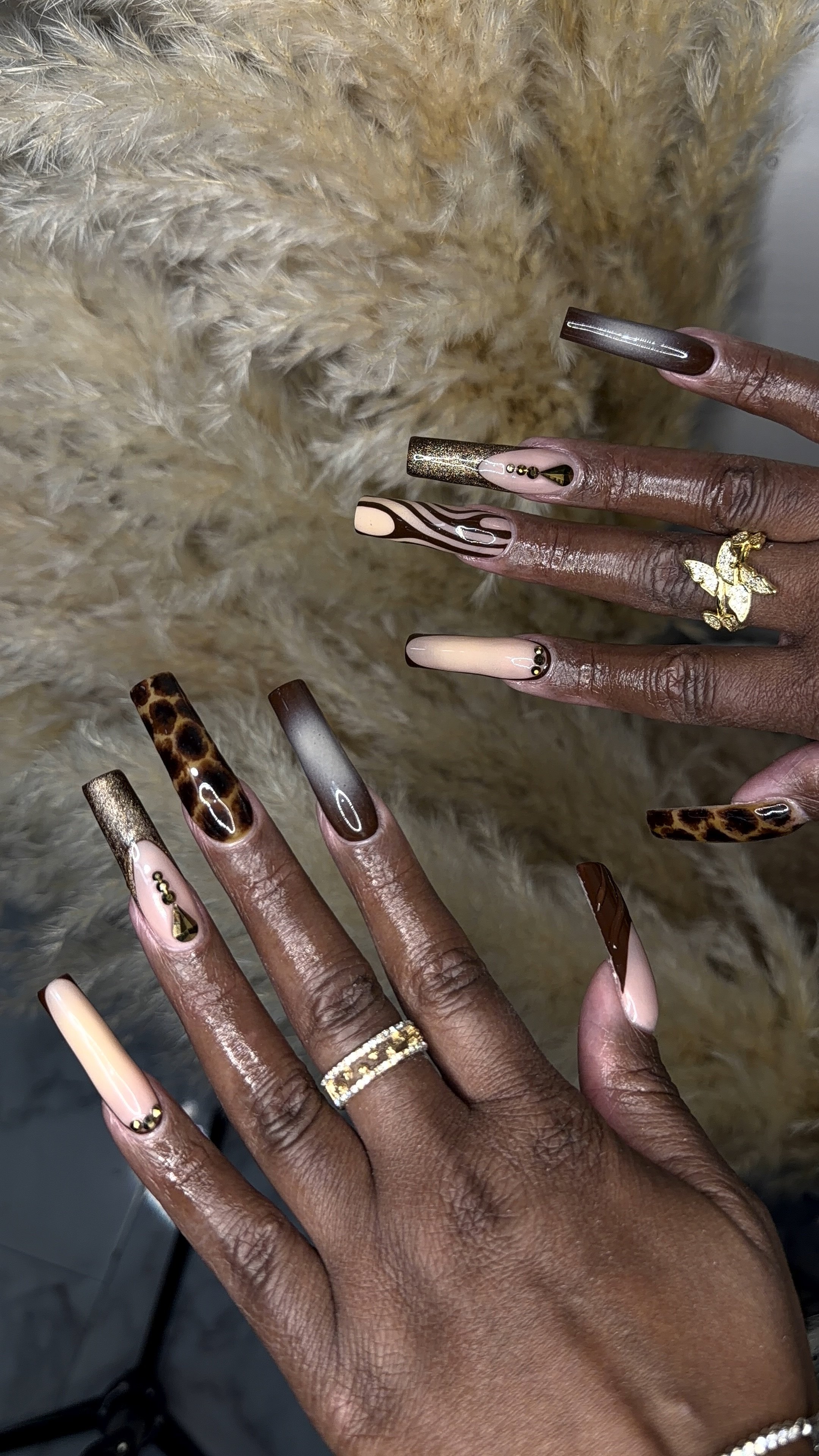 Close-up of a person's hand with long decorated nails featuring various patterns including animal print, metallics, and abstract designs, wearing multiple rings, with a furry beige background.