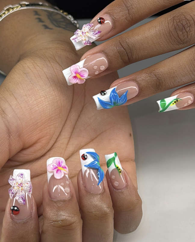 Close-up of hands with decorated nails featuring 3D floral and nature motifs, including pink, purple, and blue flowers, and green leaves on a clear base, with tiny ladybug embellishments.