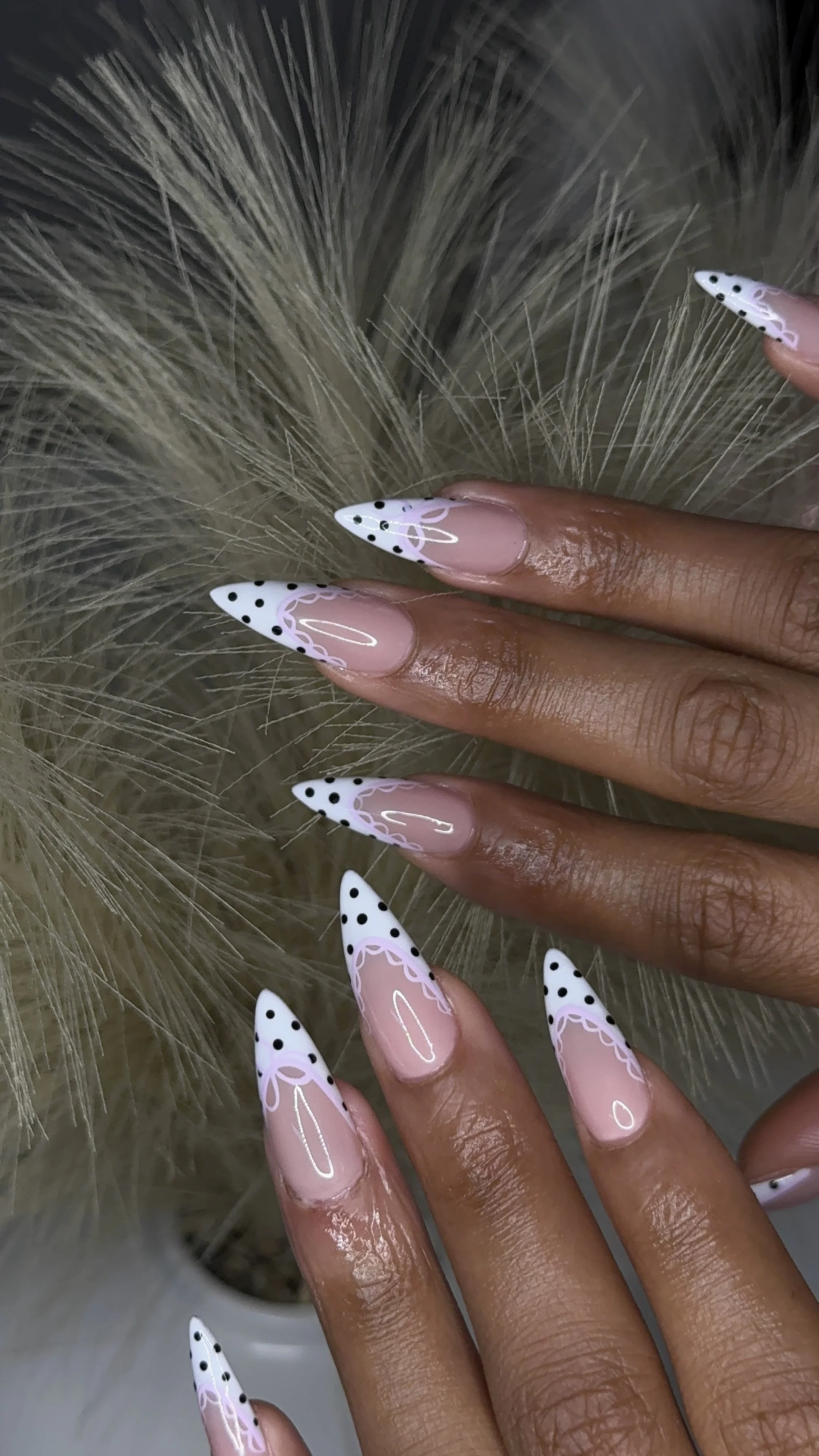 Close-up of a person's hand with long, stiletto-shaped nails featuring pink and white designs, placed against a background of light-colored fluffy plant or material.