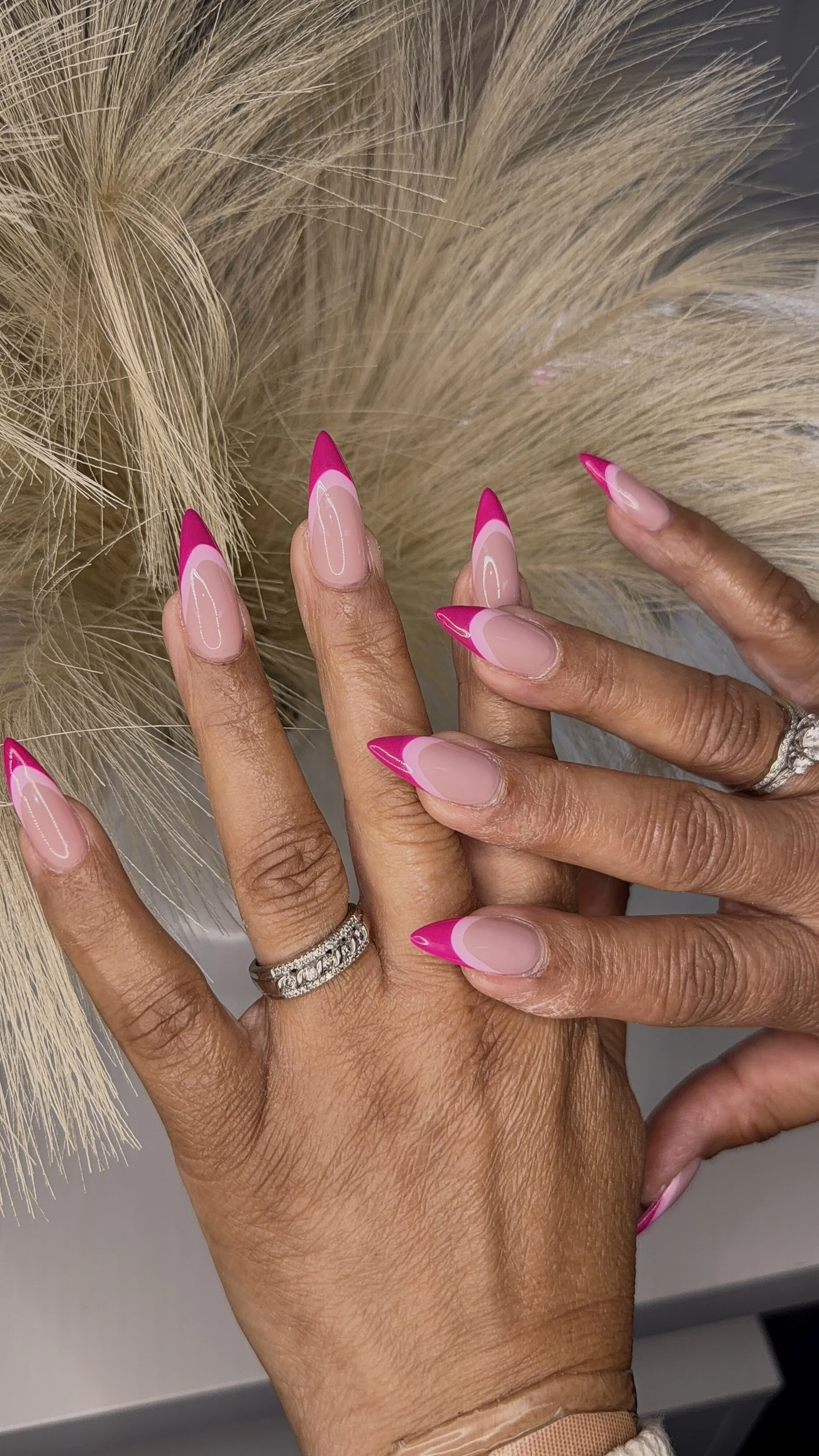 Close-up of hands with manicured nails featuring pink and nude French tips, worn with silver rings, against a background of blonde hair.