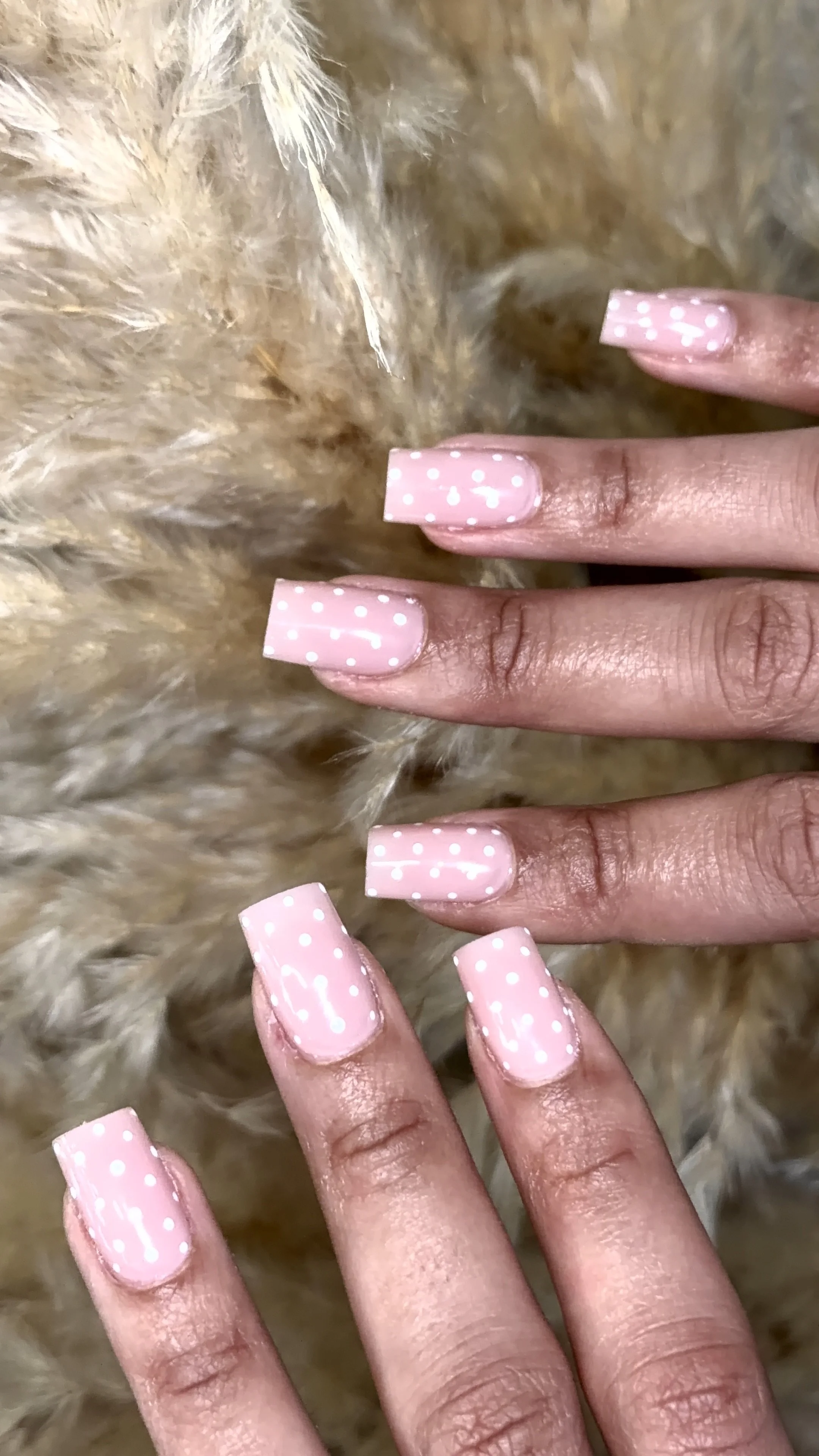 Close-up of a hand with pink and white polka dot painted nails resting on a furry, light tan textured surface.