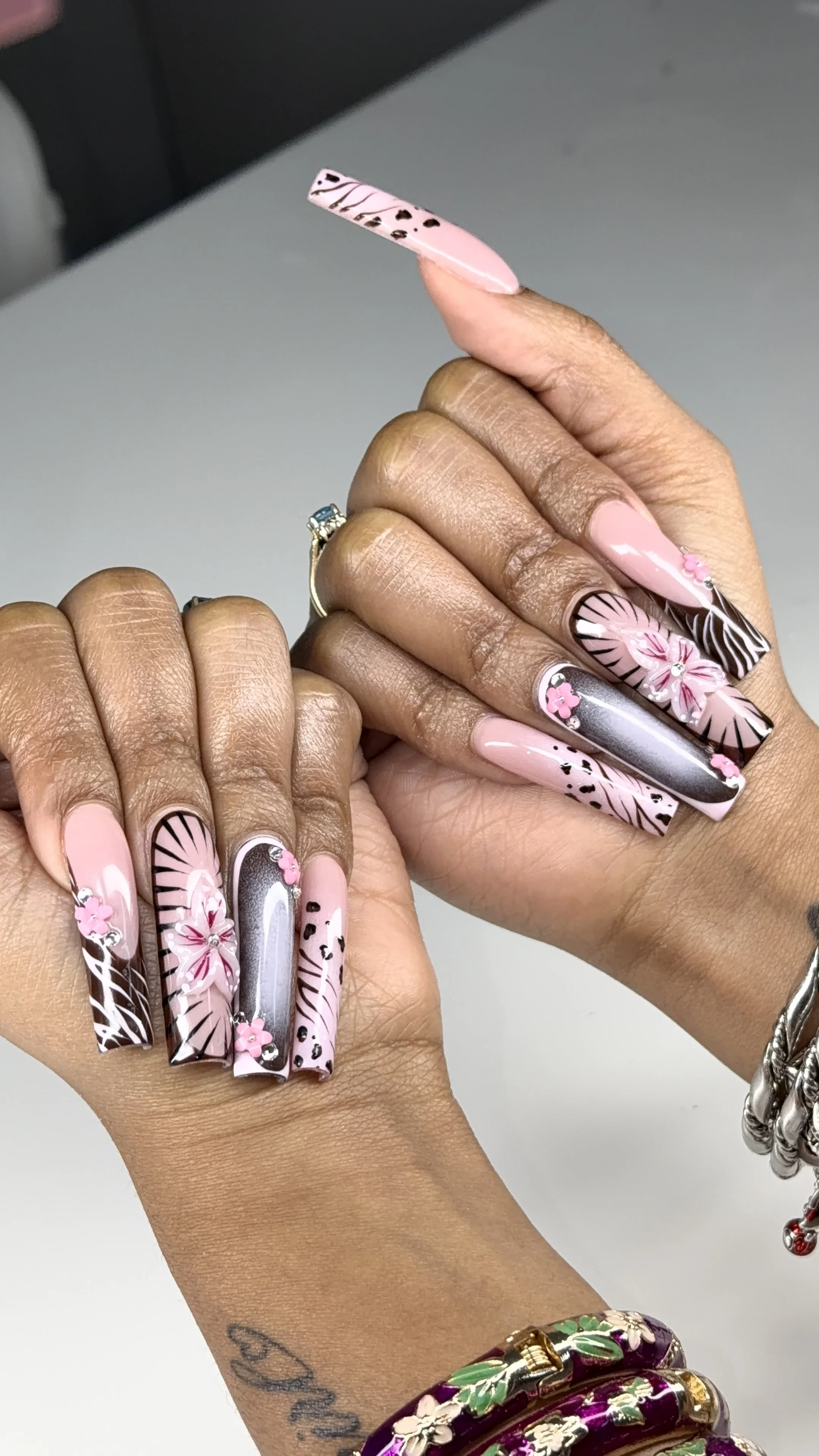 Close-up of a person's hands showing decorated long acrylic nails with pink, black, white, silver, and floral designs, including rhinestones and 3D flowers.
