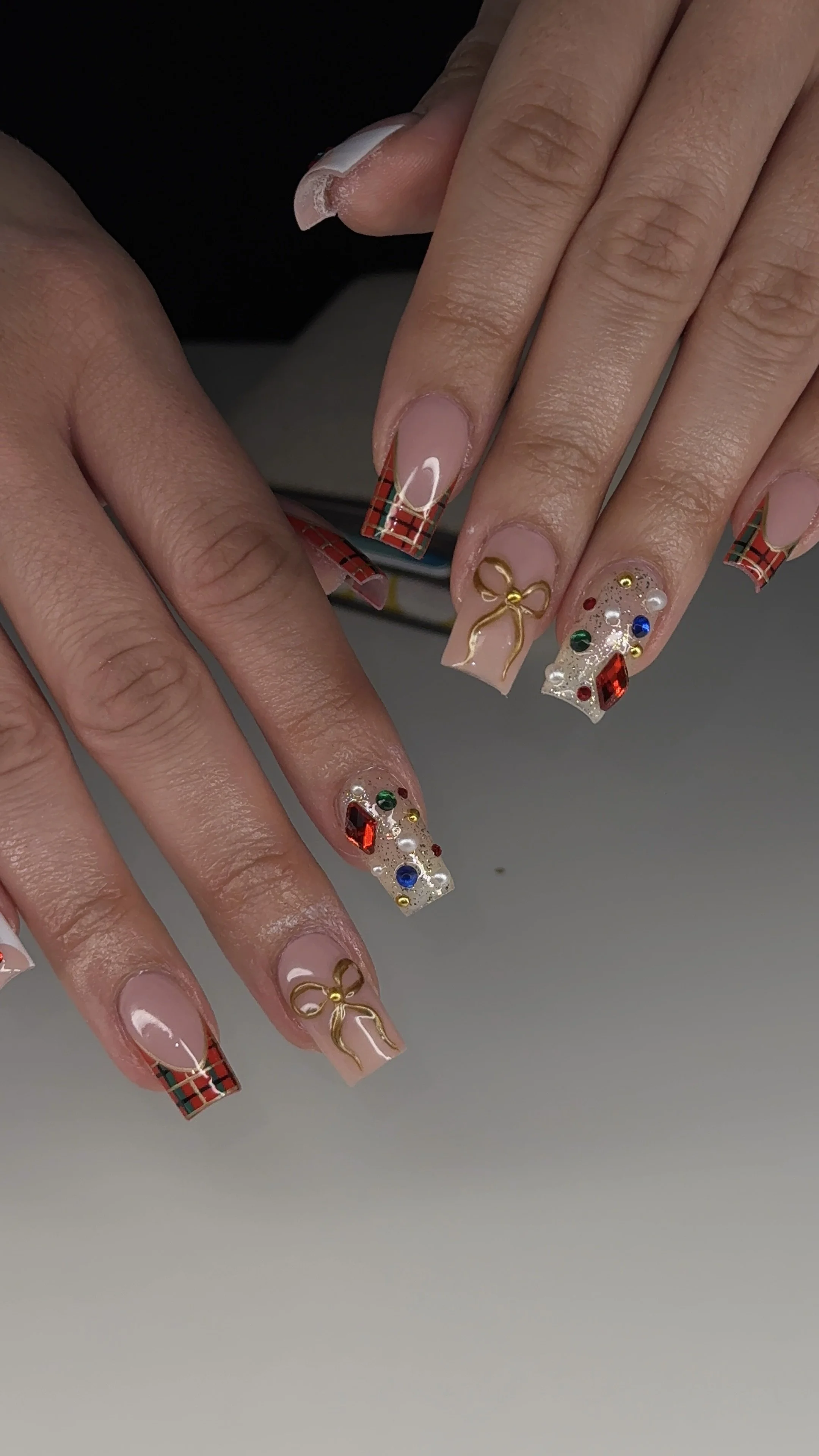 Close-up of decorated Christmas-themed manicured nails featuring plaid patterns, gold bows, and colorful rhinestones.