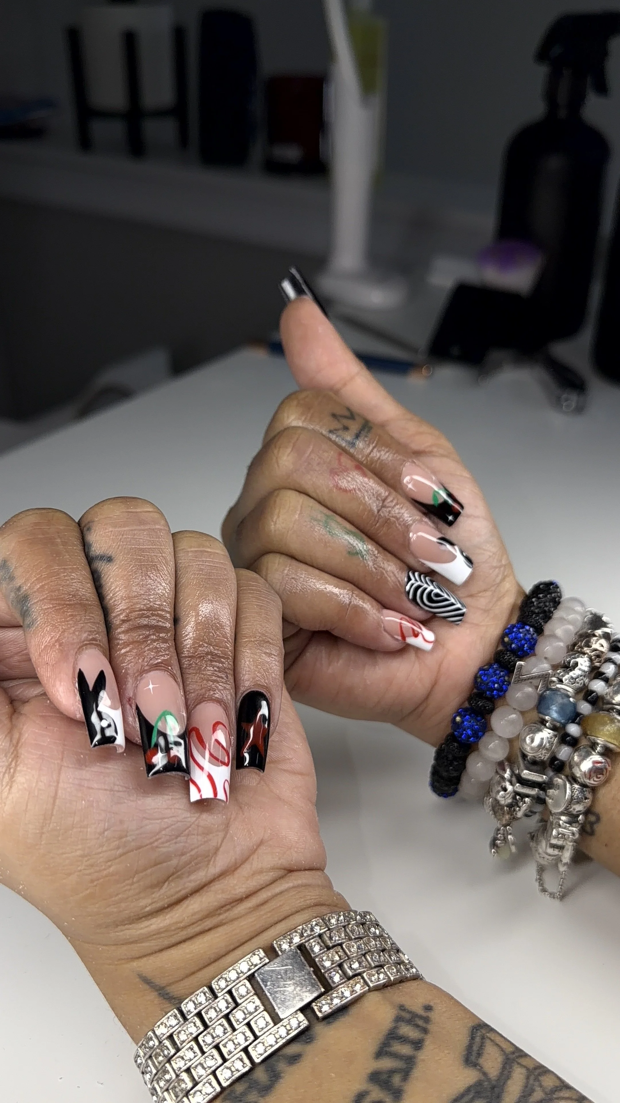 Close-up of a person's hands with decorated Christmas-themed nails, jewelry including a sparkling bracelet, and multiple bracelets, resting on a white surface with a blurred background.
