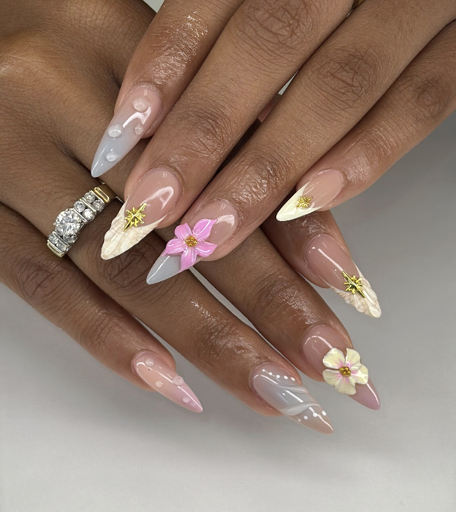 Close-up of hands with long stylish nails featuring pastel pink, white, and gray colors, decorated with 3D flowers, gold accents, and rhinestones.