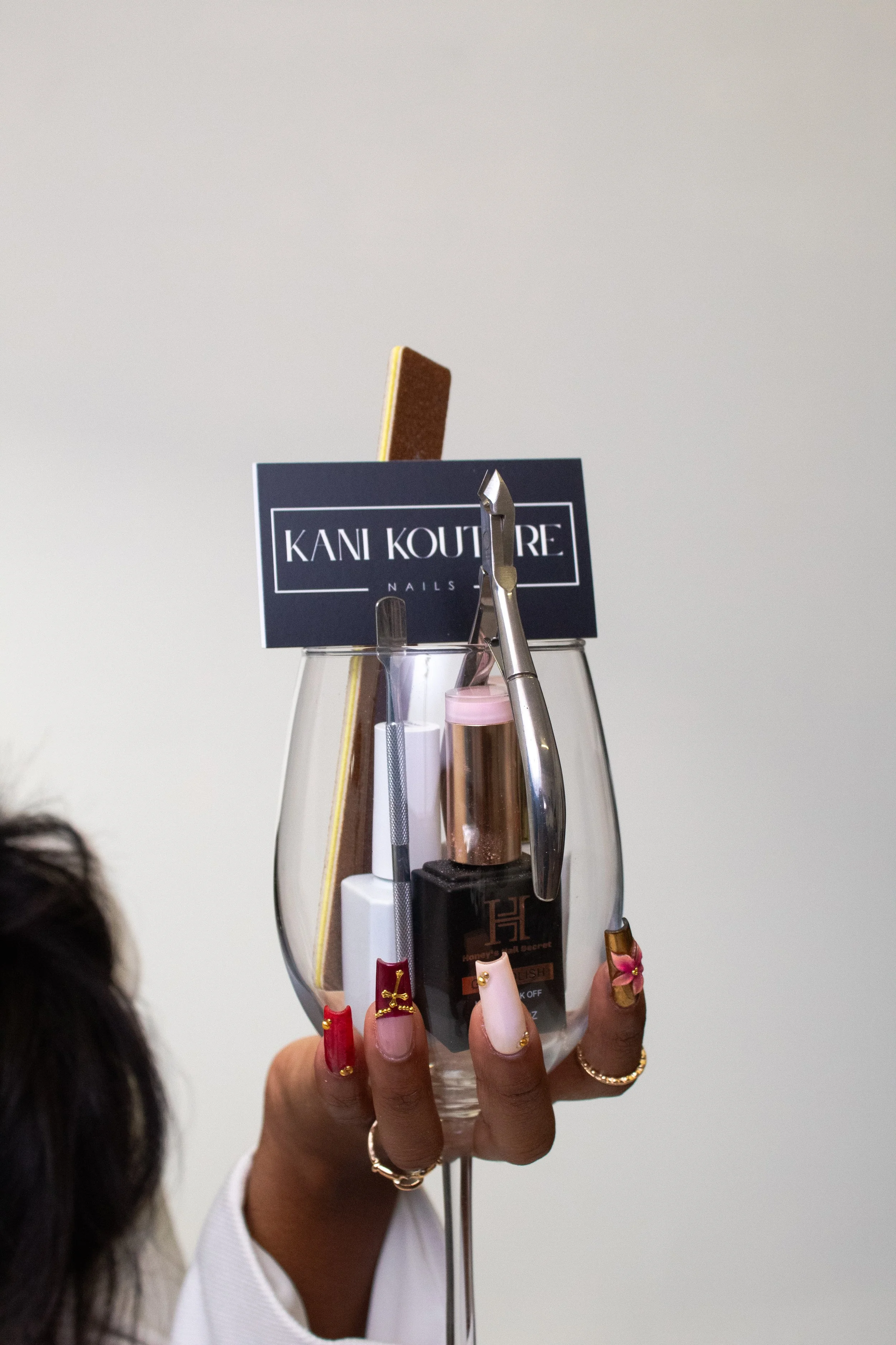 Hand holding a wine glass filled with nail and beauty tools, including nail files, a cuticle pusher, and a bottle of nail polish, with a business card that reads "KANI KOUTURE".