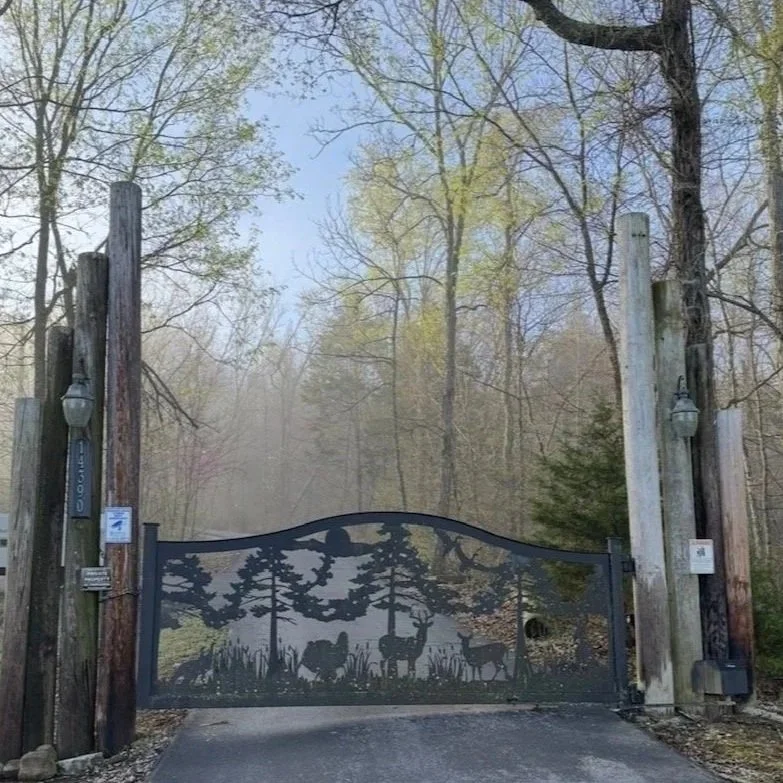 A decorative black metal gate featuring a silhouette design of forest animals, trees, and a moon, flanked by wooden posts with lanterns, set in a wooded area.