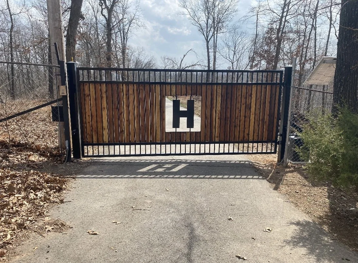 A black metal gate with a wooden panel and a large letter H in the center, blocking a driveway in a wooded area.