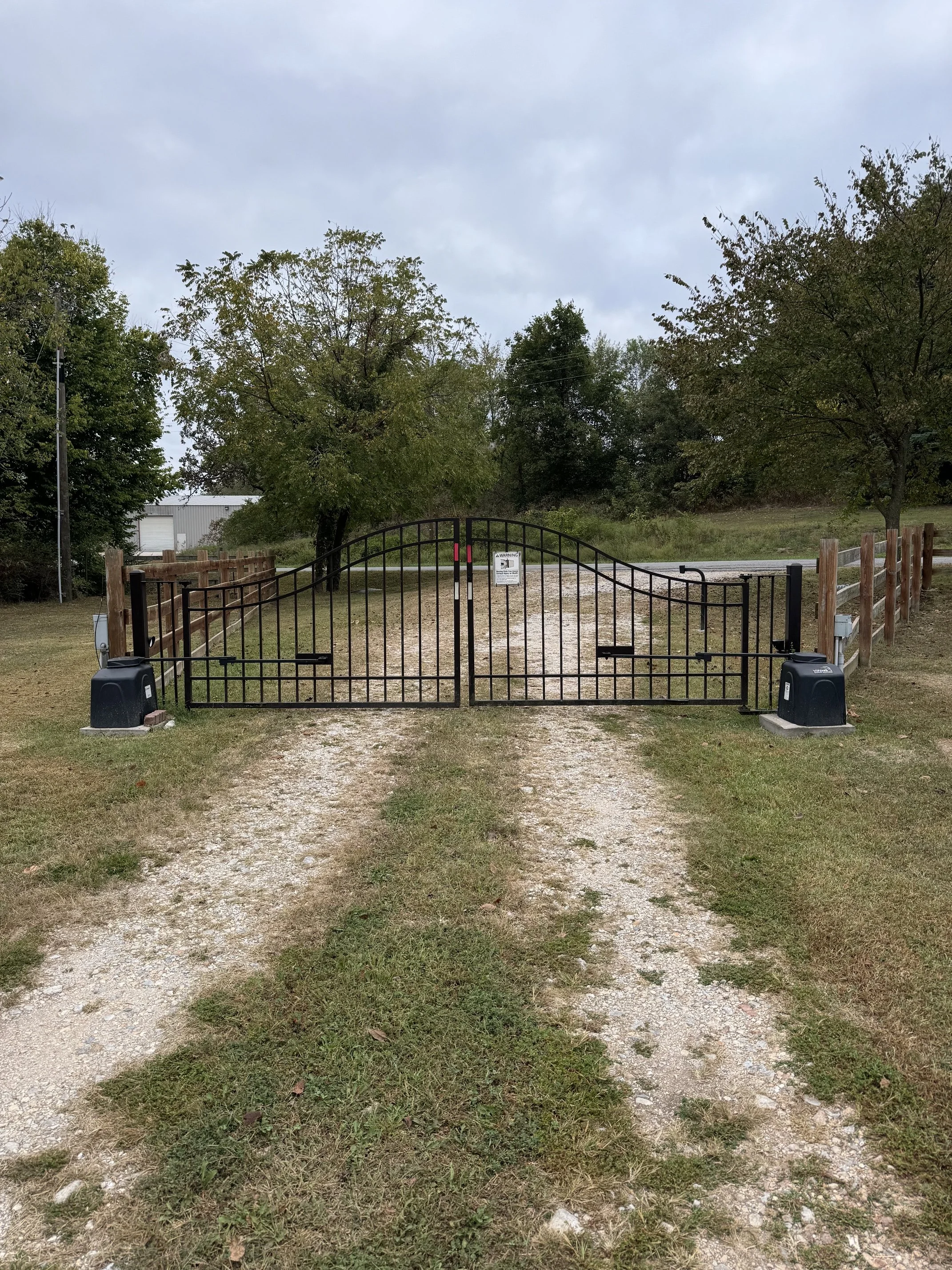 A black metal gate situated on a dirt and grass pathway, flanked by a wooden fence on both sides, with trees in the background and a partly cloudy sky overhead.