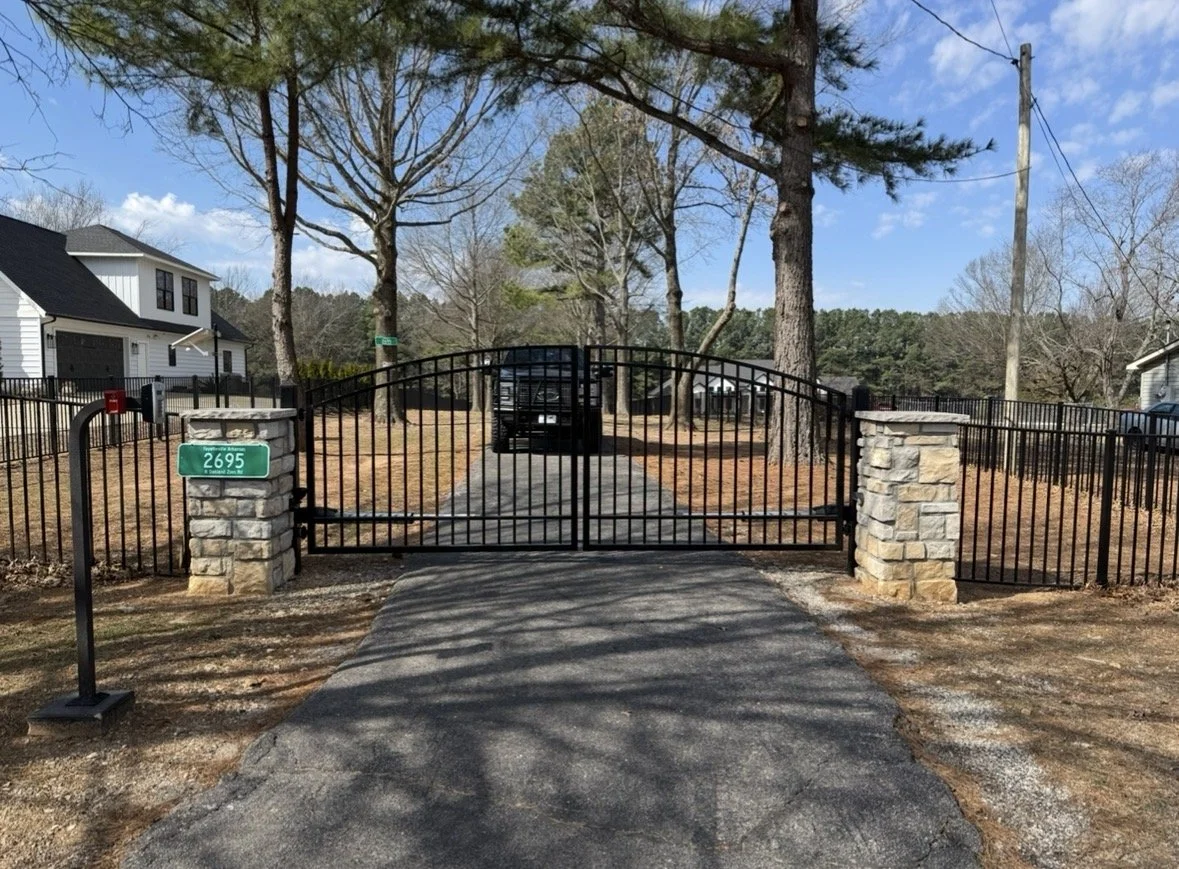 Black metal gate with stone pillars at entrance of residential property, driveway and trees in background, blue sky with clouds.