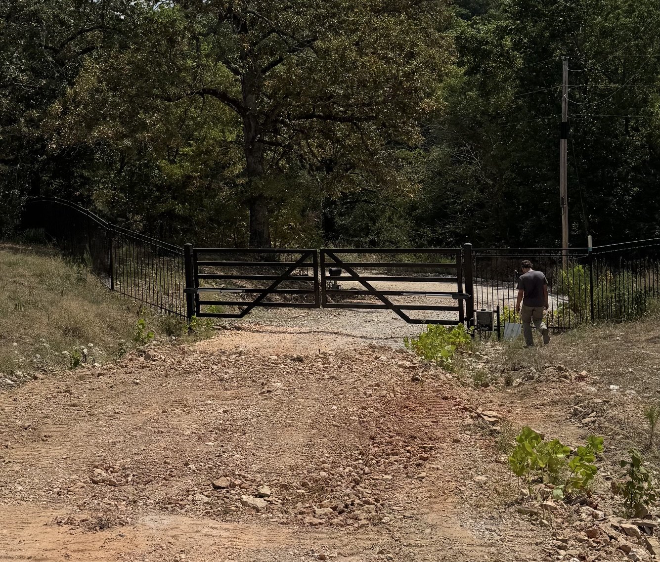 A man walking through a black metal gate in a rural area with trees and utility pole in the background.
