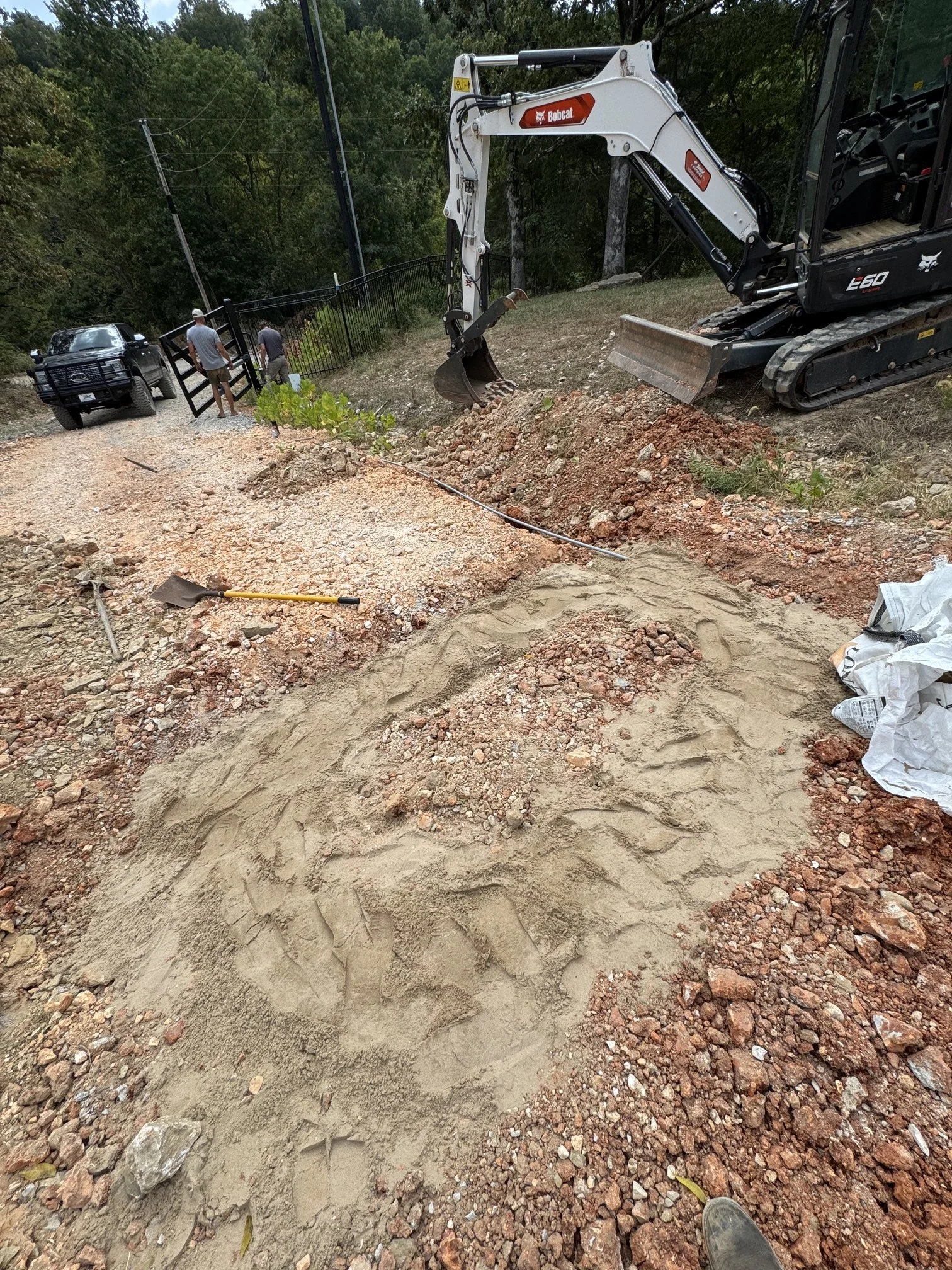 Excavation site with a small tracked excavator digging a trench, a pile of dirt, and two workers walking near a black truck in a wooded area.