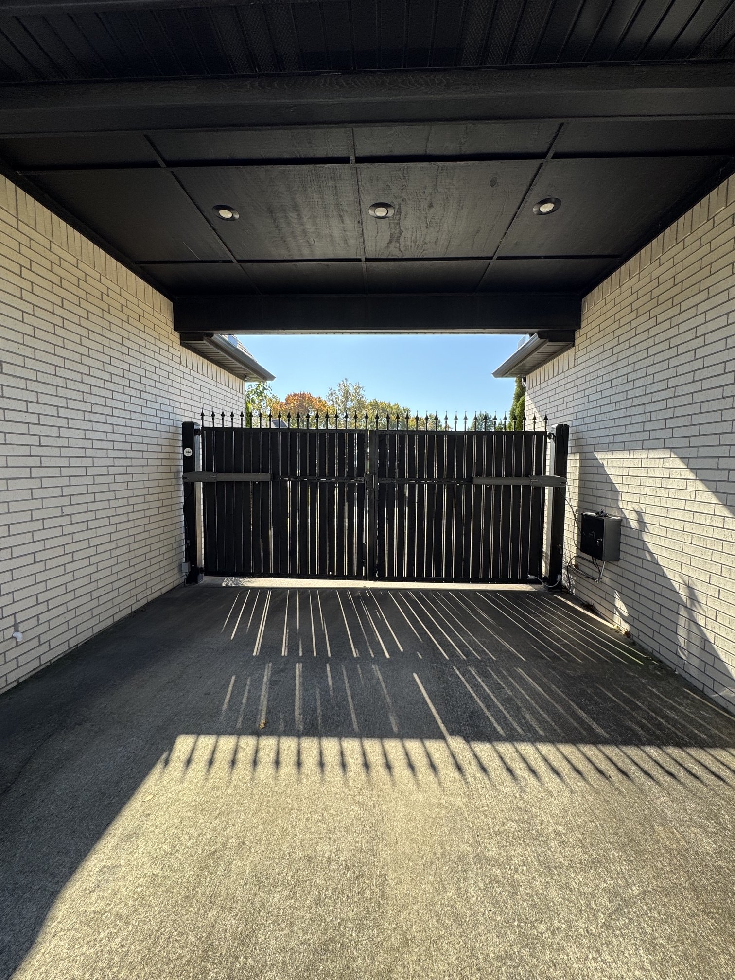 View of a garage with a black gate, brick walls on either side, and a ceiling with embedded lights. Shadows of the gate are cast onto the concrete floor, with sunlight illuminating part of the floor and walls.